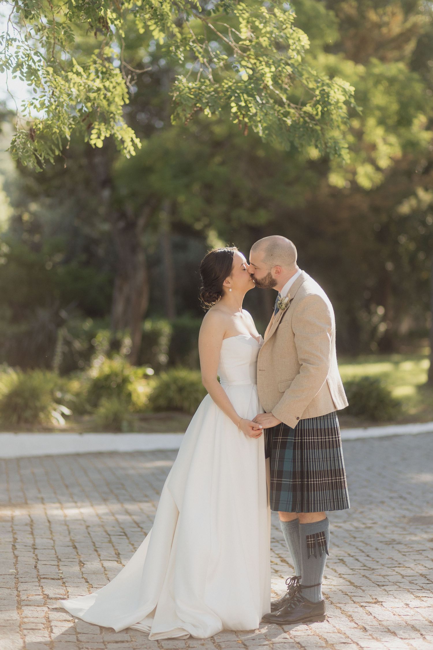 Couple shares intimate moment on cobblestone path surrounded by green trees.
