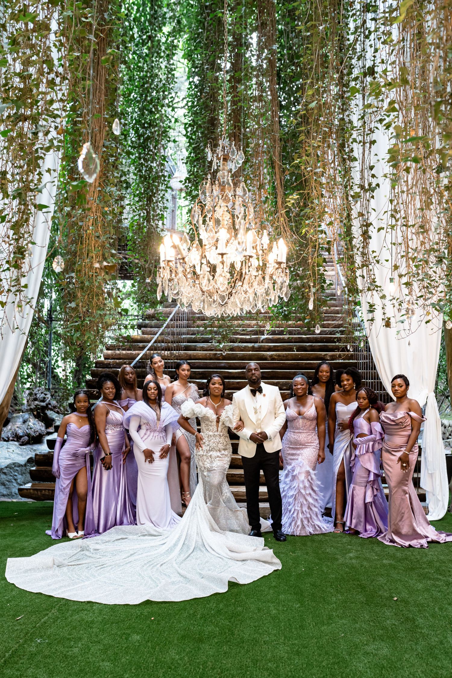 Wedding party poses for photos on a lawn with hanging greenery and white draping.