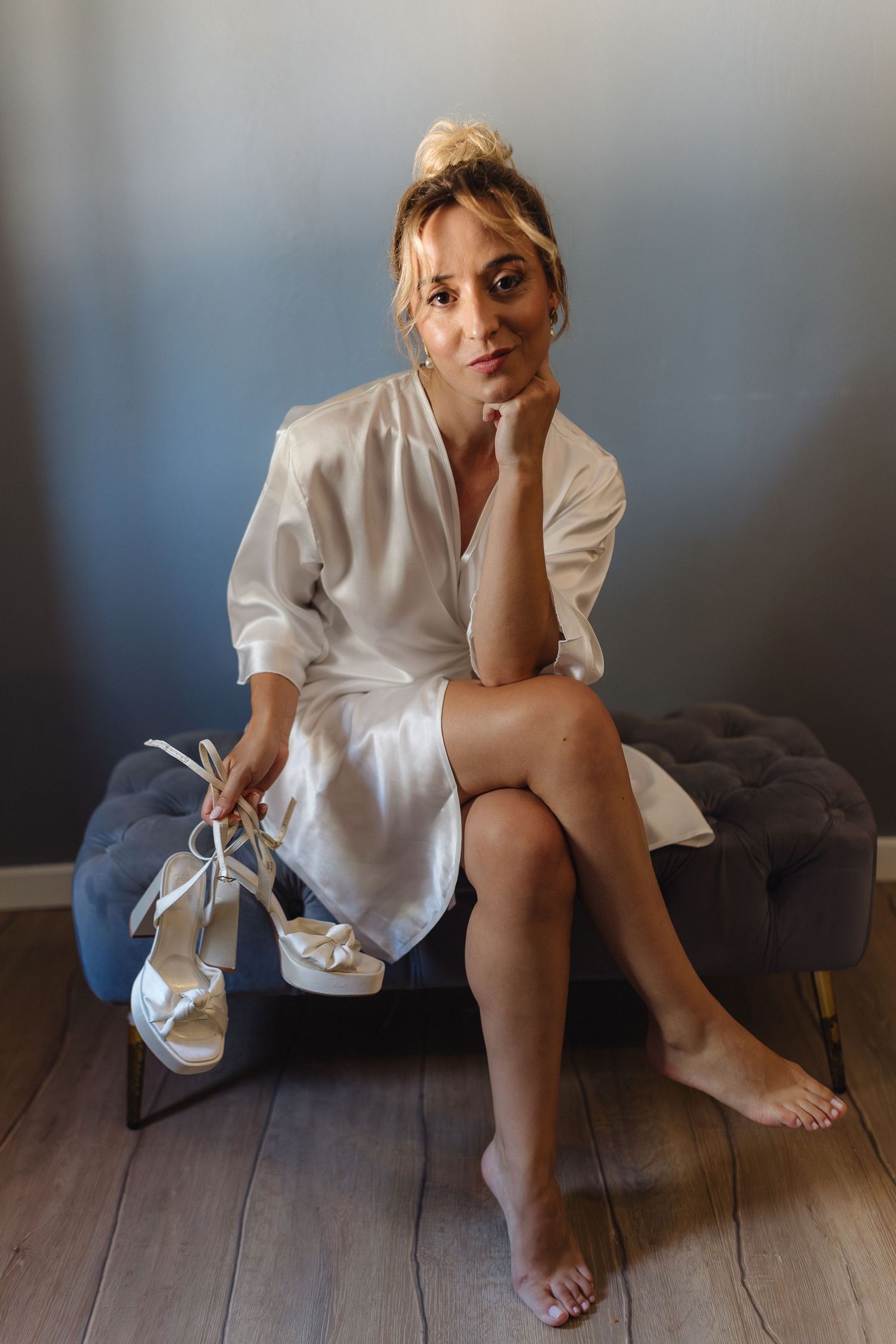 Woman in white oversized shirt poses casually on wooden floor in minimalist interior setting.