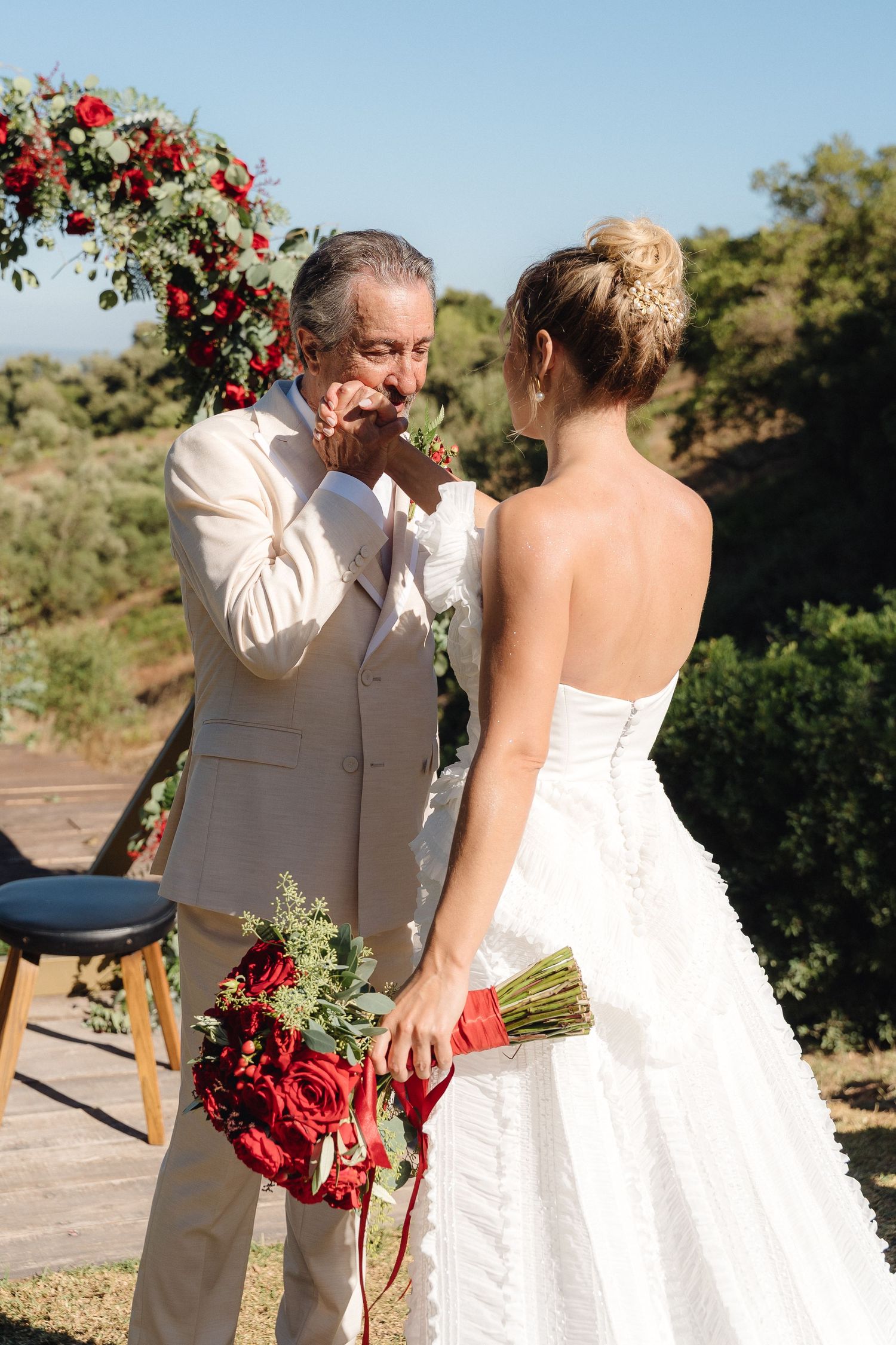 Bride in strapless gown and guest in cream suit share touching interaction with red roses in background.