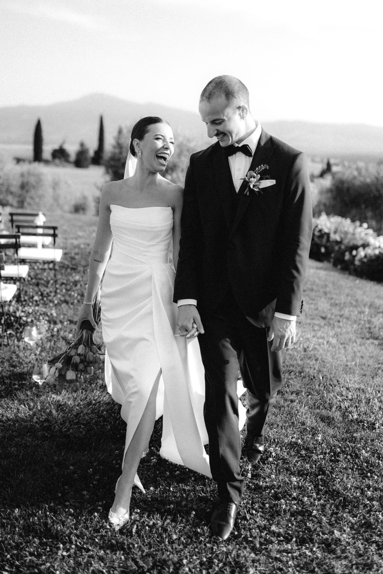 Happy bride and groom walk hand in hand laughing after their outdoor wedding ceremony in the Tuscan countryside.