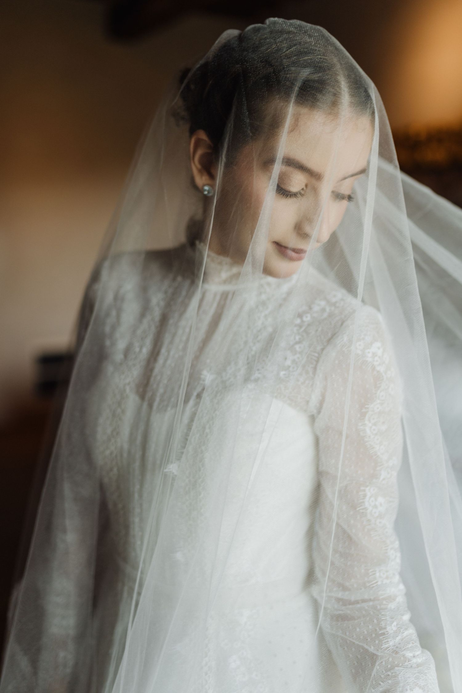 Elegant bride in long-sleeve lace wedding dress with sheer veil draped over her head, eyes closed, soft natural light.
