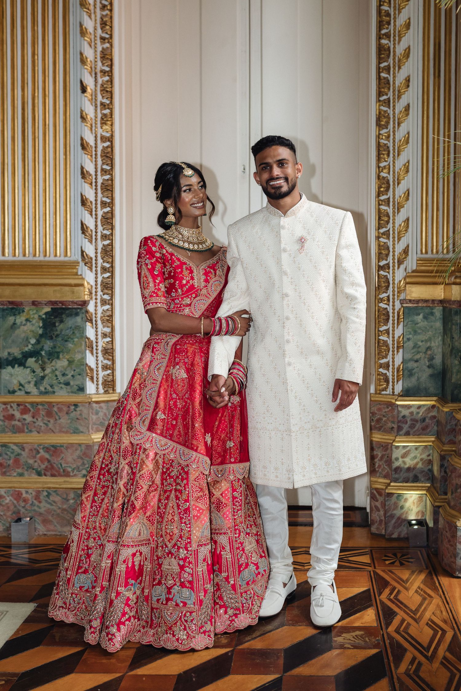 Traditional wedding attire featuring red embroidered lehenga and white sherwani in elegant room.