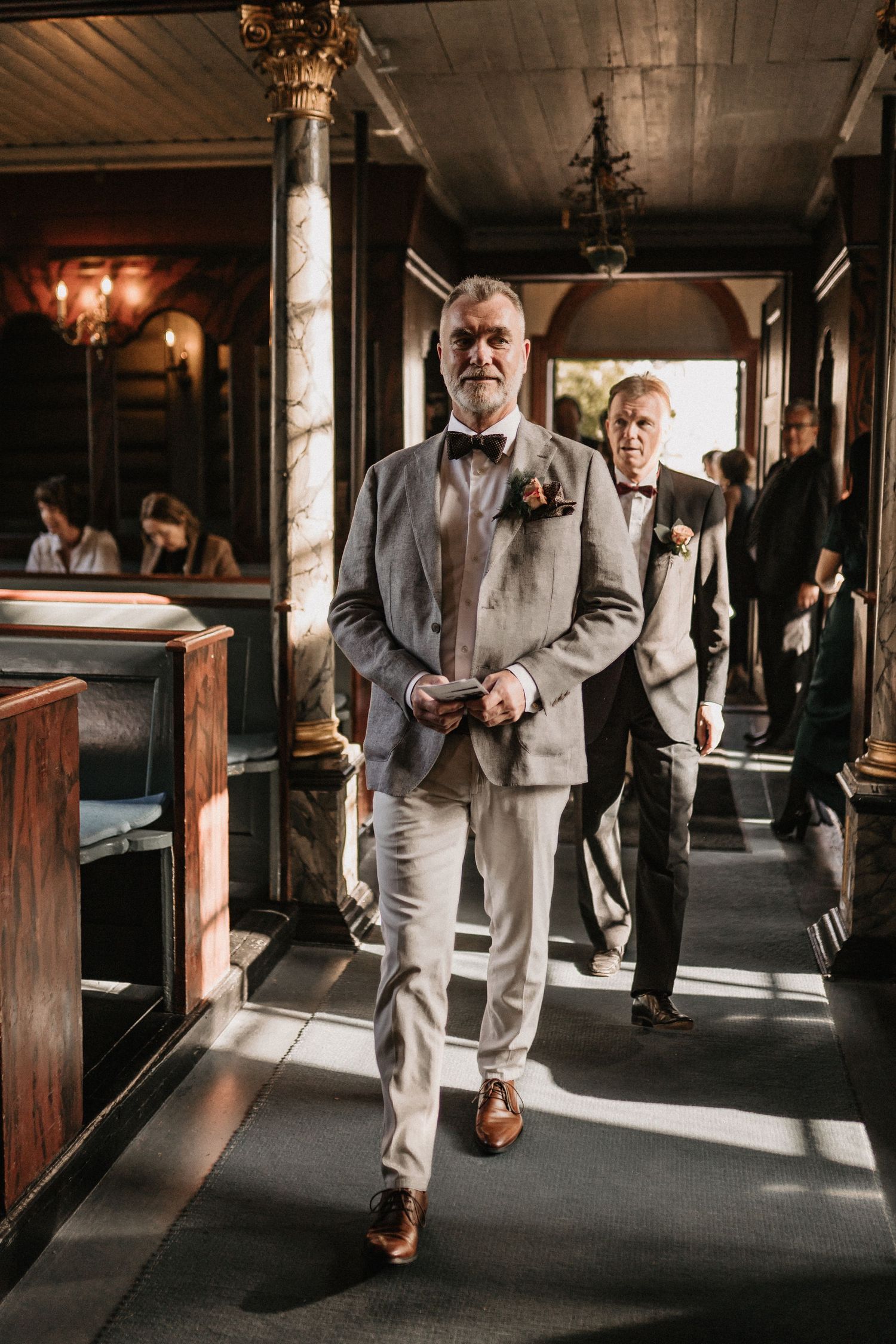 Wedding guests in formal attire walking through a vintage wooden corridor with warm lighting.