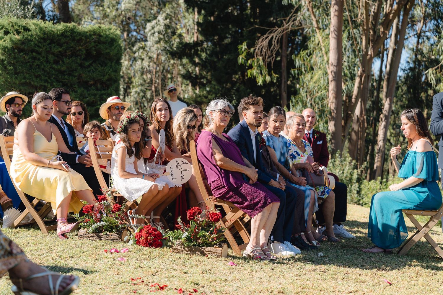 Wedding guests seated on wooden chairs during outdoor ceremony surrounded by trees.