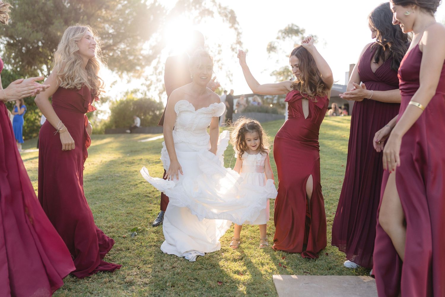 Wedding party in burgundy dresses dancing outdoors in golden sunlight.
