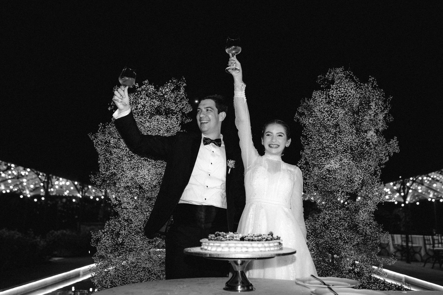 Black and white photo of a bride and groom toasting with champagne glasses at their elegant outdoor wedding reception.