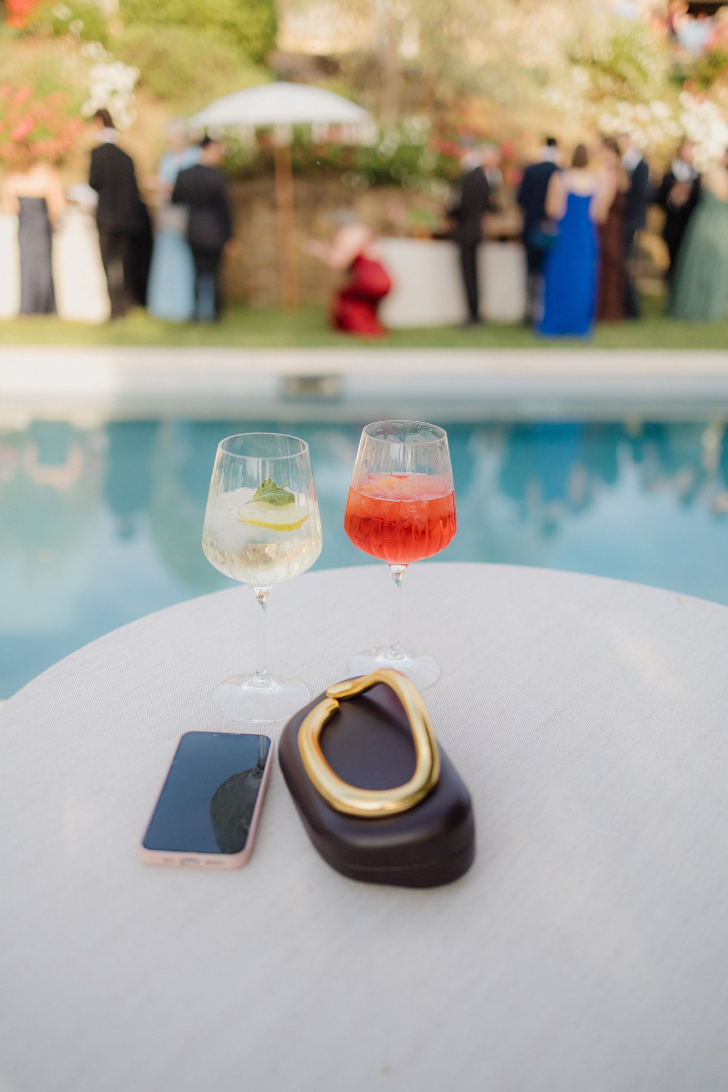Two cocktail glasses beside a smartphone and clutch purse on a white table near a wedding reception pool.