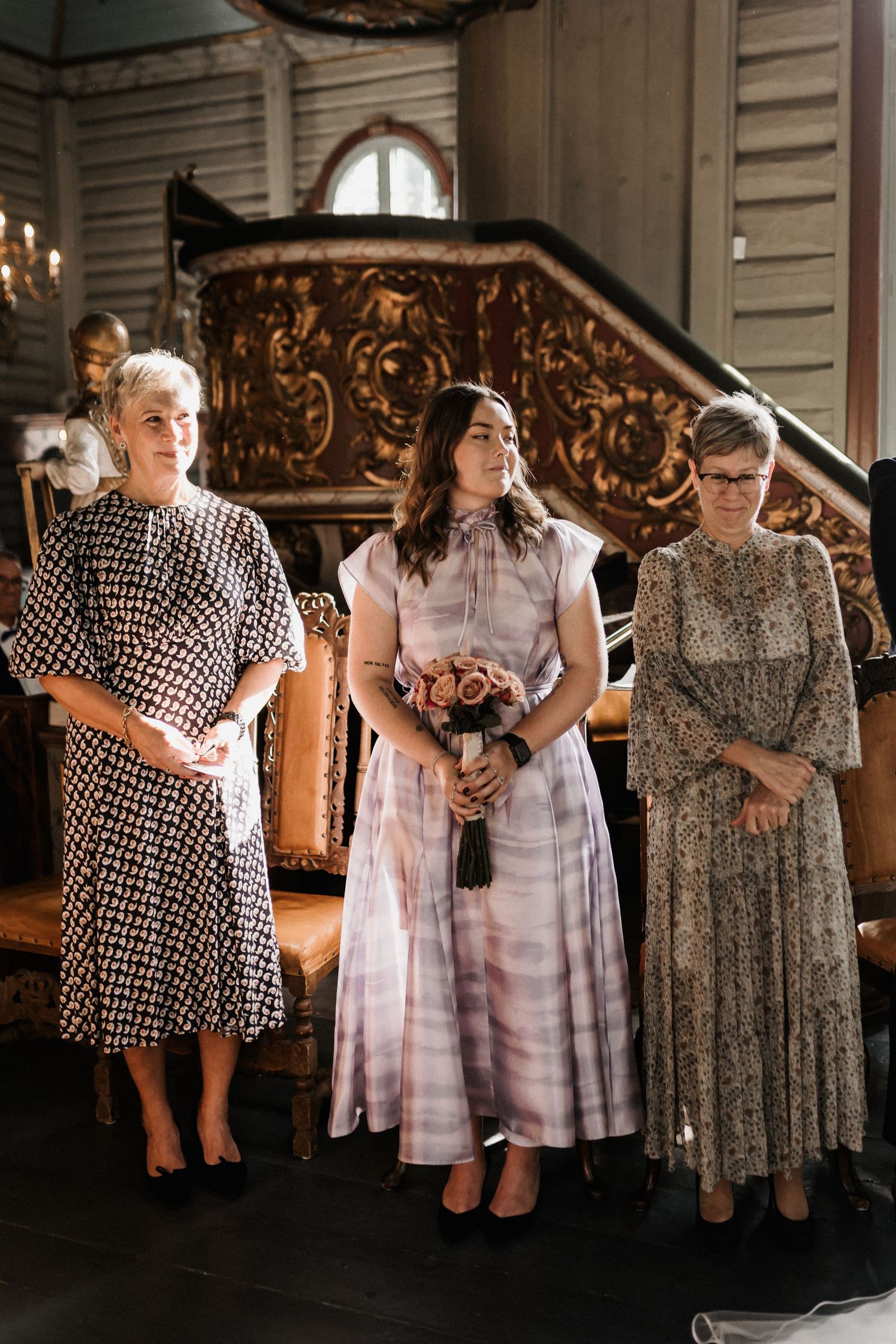 Three guests in patterned dresses stand together during a wedding ceremony in an ornate church setting.