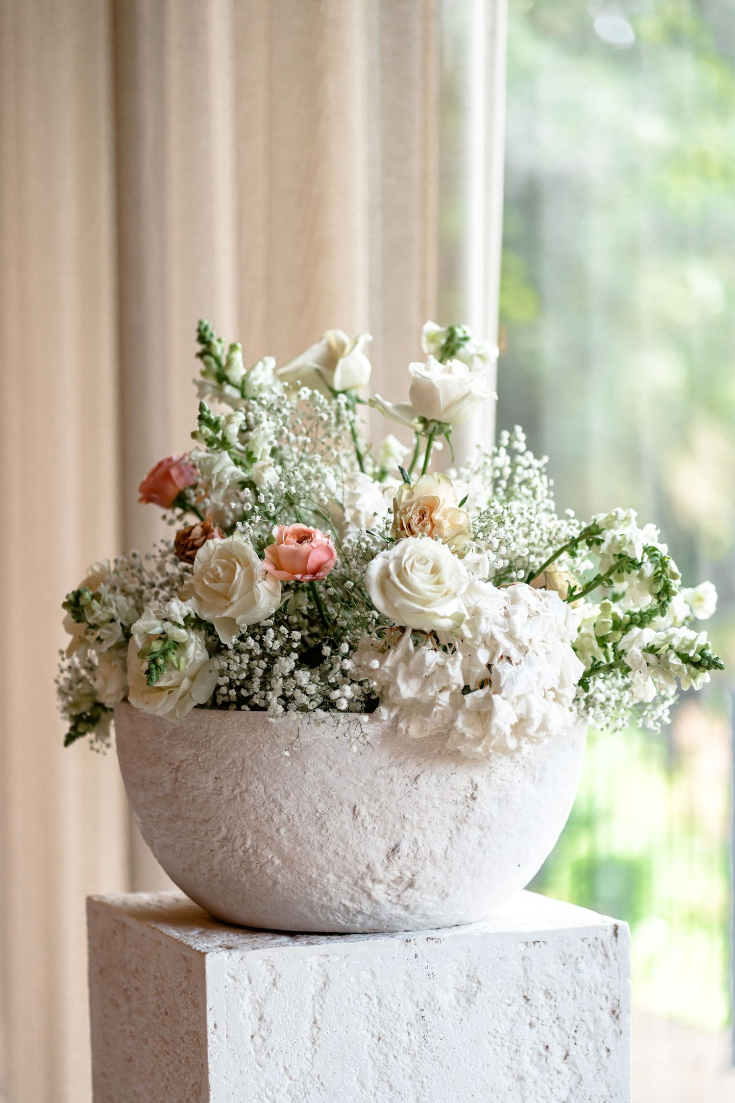 Delicate white and peach floral arrangements in textured stone bowls against a light curtained backdrop.