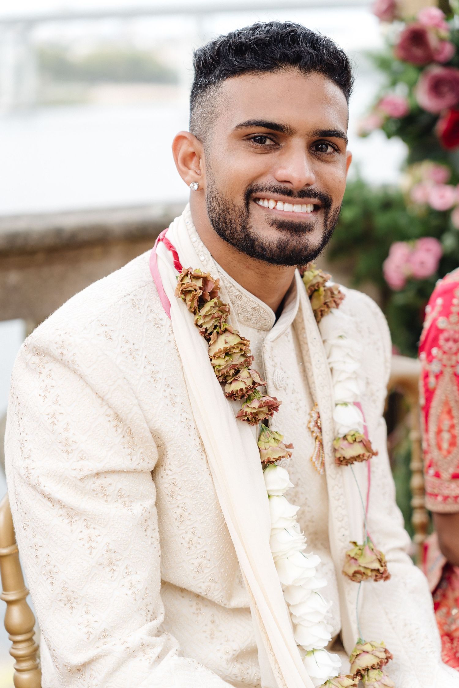 Groom wearing cream sherwani and floral garland at outdoor Indian wedding celebration.