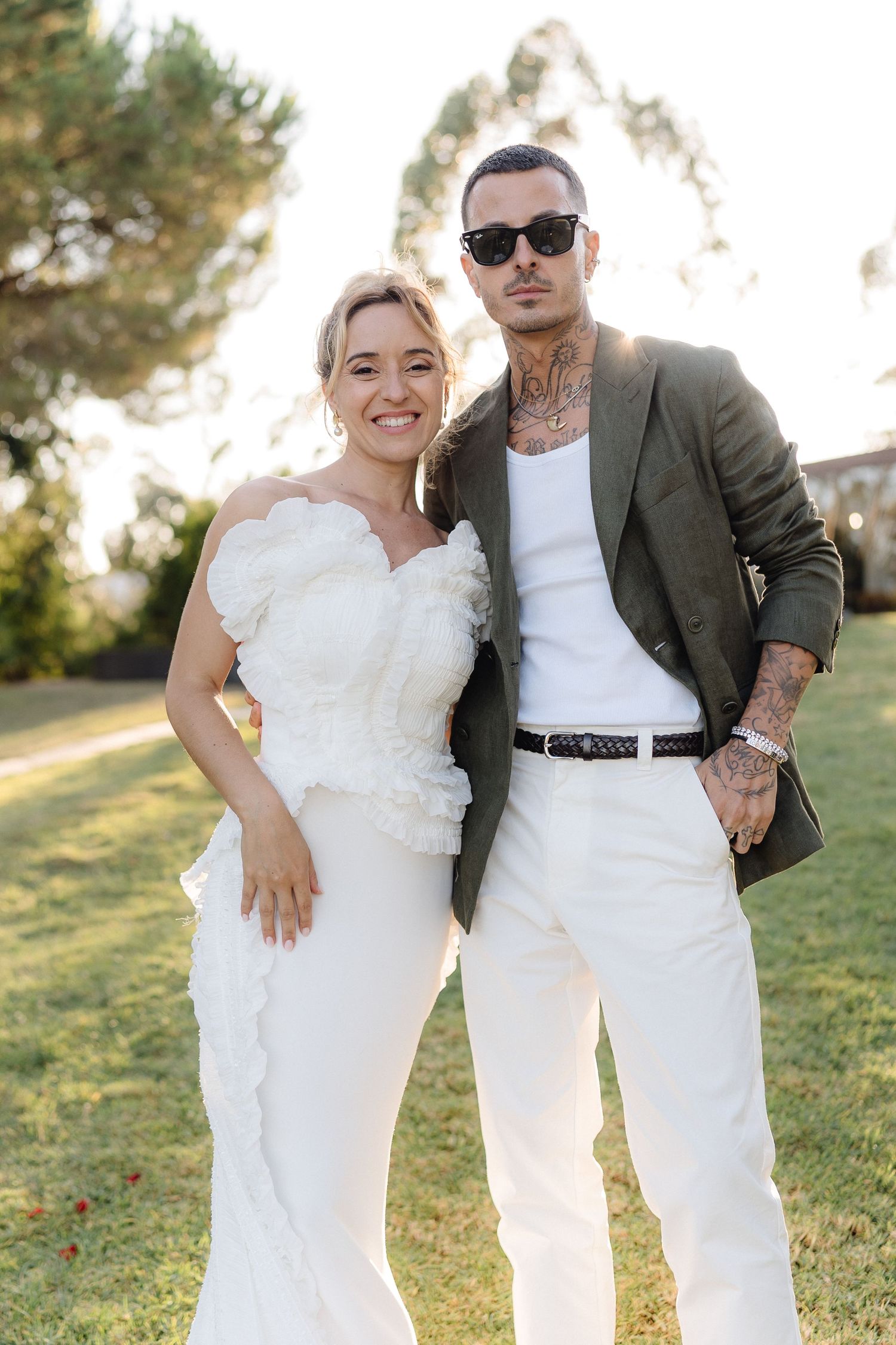 Wedding couple poses together in white attire against a sunlit outdoor backdrop.