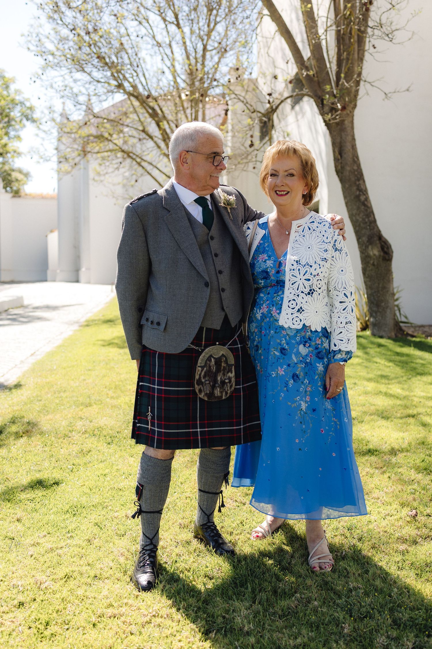 A couple in formal attire, including a traditional Scottish kilt and blue dress, pose together outdoors.