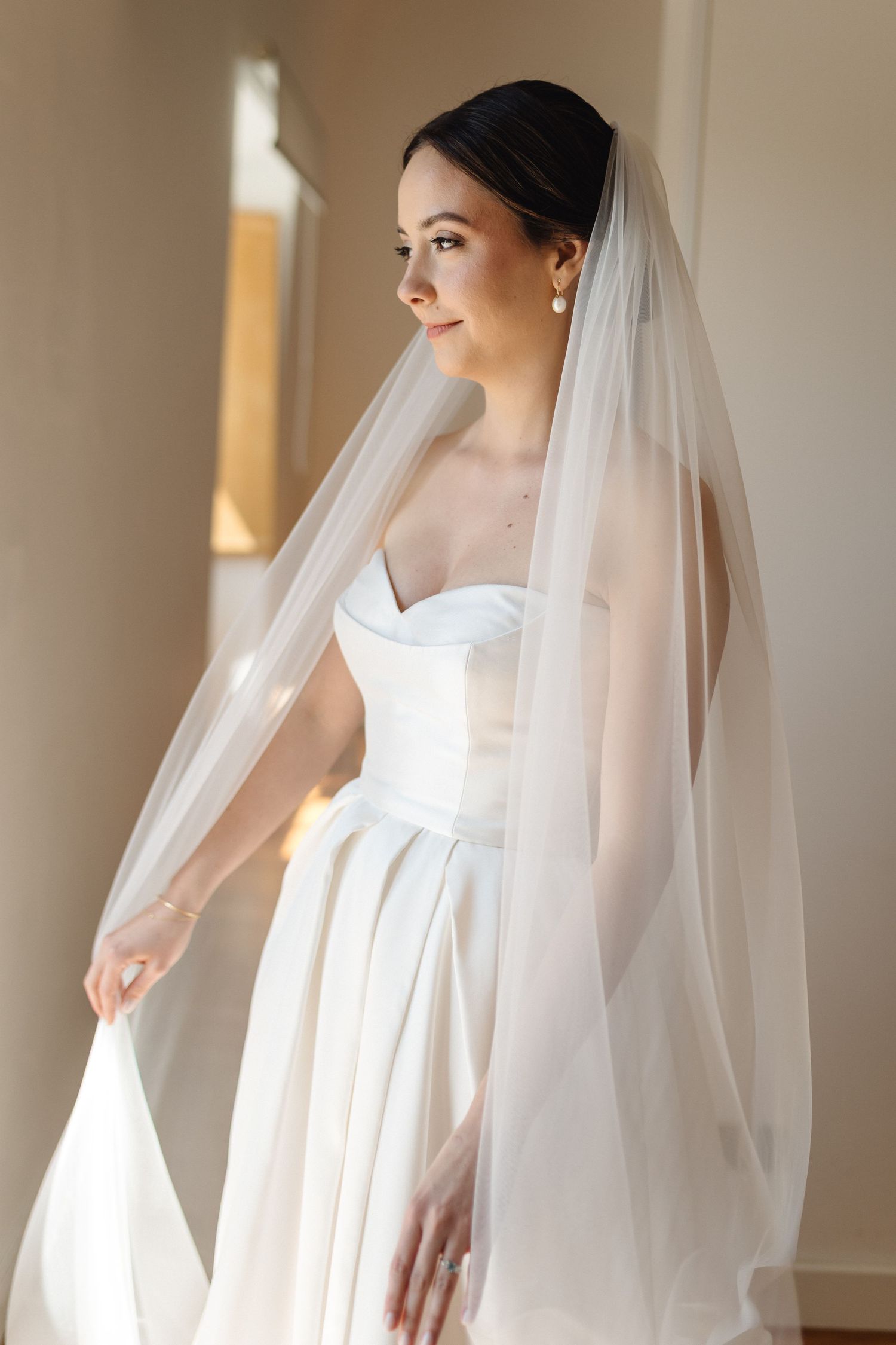 Bride in classic white wedding dress and veil standing in sunlit hallway.