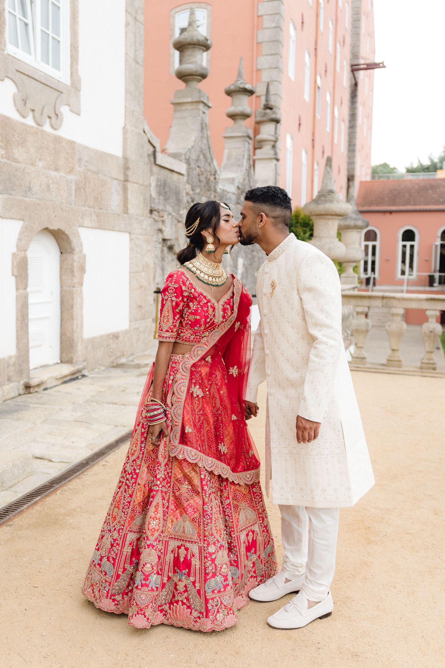 Couple in traditional red lehenga and white sherwani pose at elegant venue.