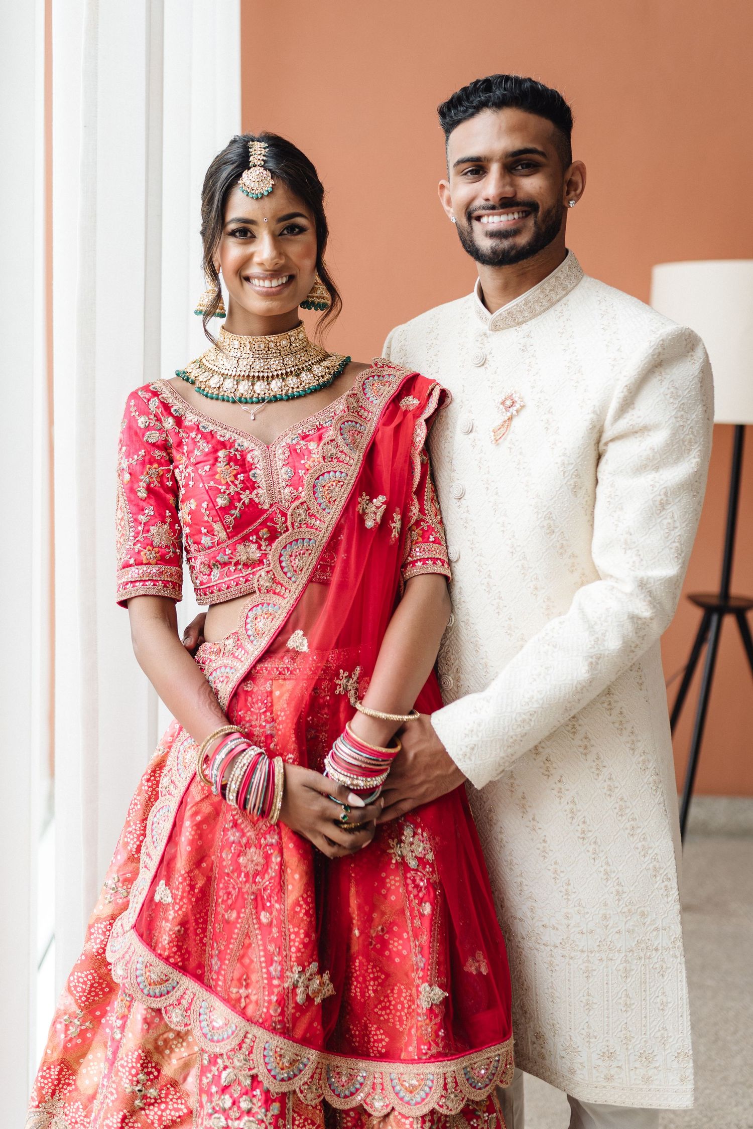 Wedding couple in traditional Indian attire with red lehenga and white sherwani against coral wall.
