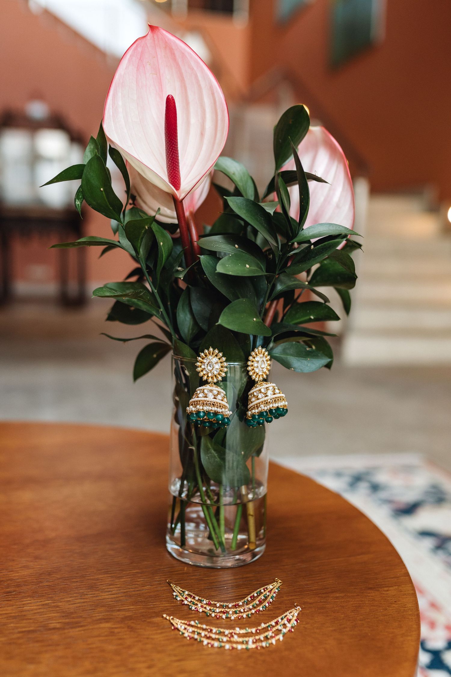 Elegant jewelry and pink anthurium flowers displayed in a glass vase on wooden table.