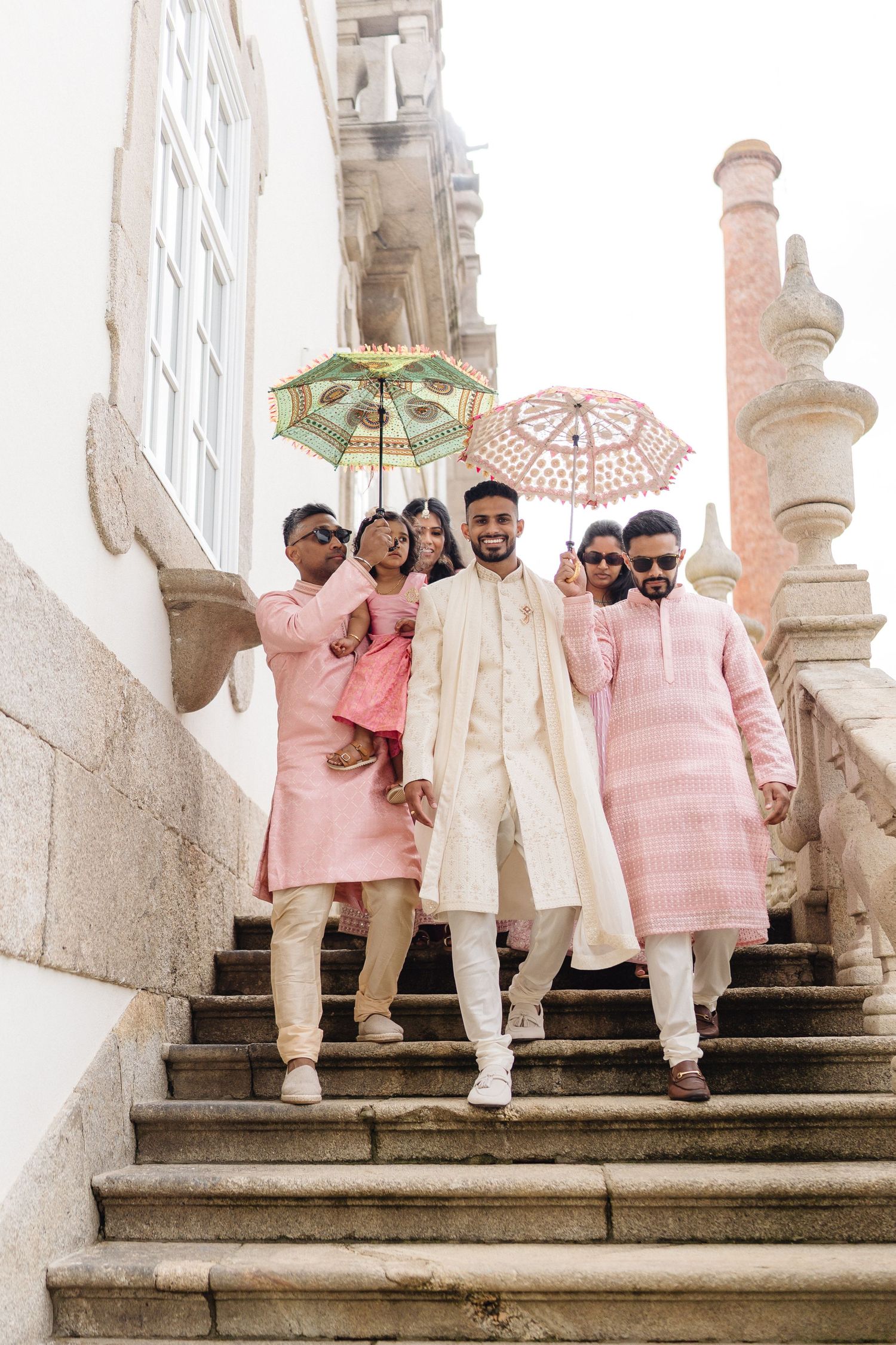 Wedding party in coordinated pink and cream outfits posing on stone steps with parasols.