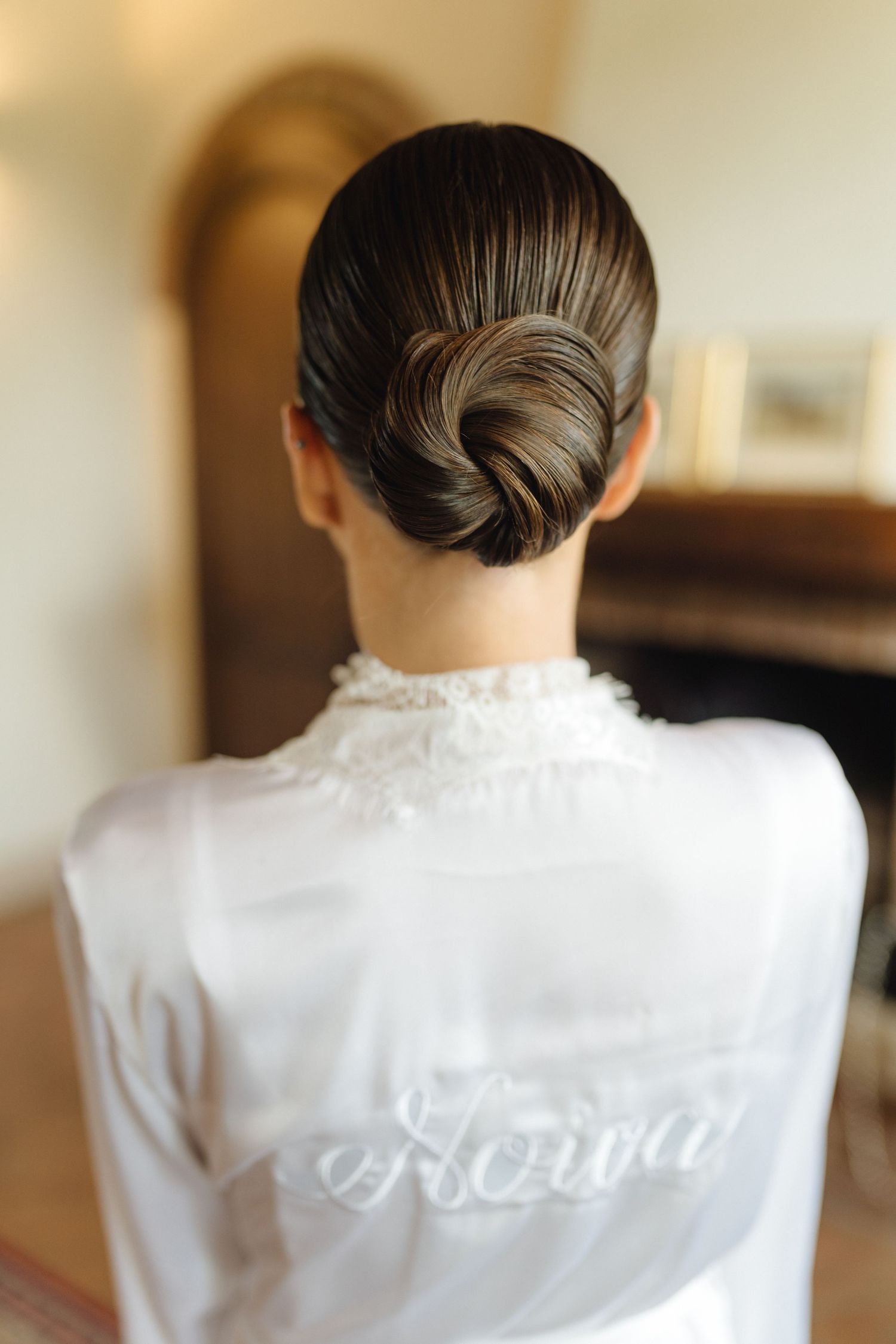 Bride with elegant low chignon bun wearing white satin robe with Noiva script on back.