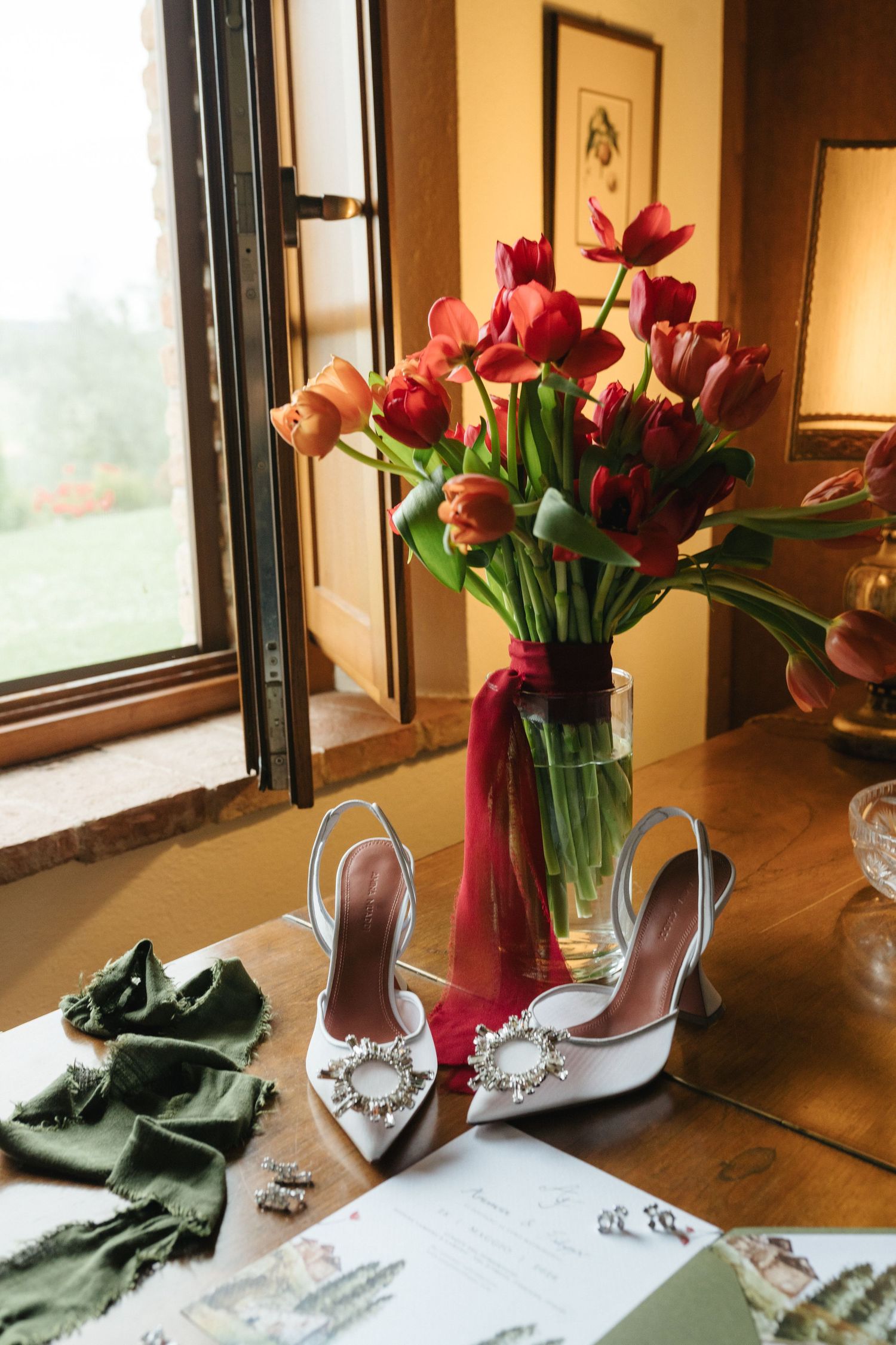 White bridal heels with crystal buckles beside red tulips bouquet and green accessories on wooden table near window.
