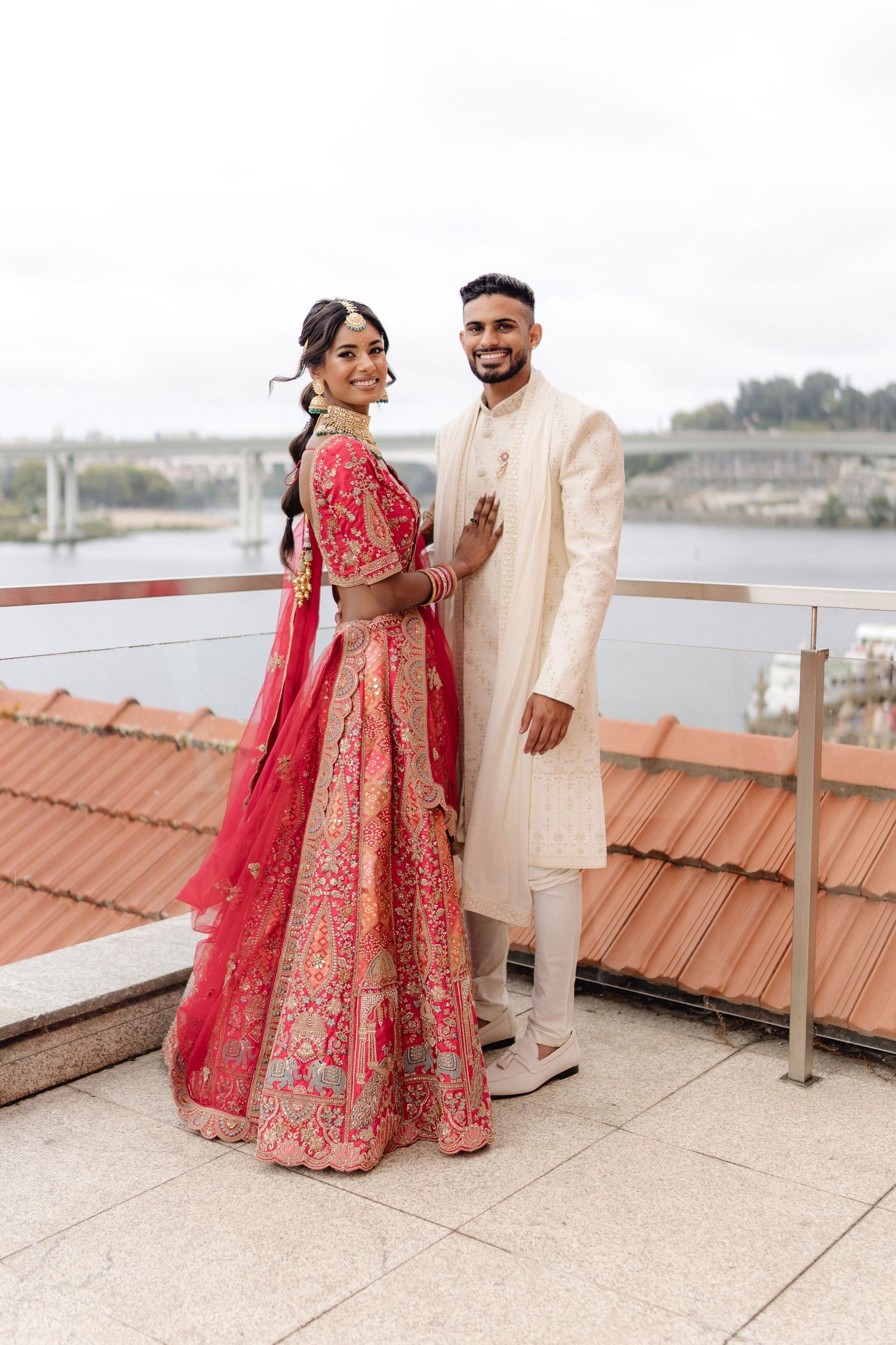 Traditional Indian wedding attire featuring red and gold lehenga and cream sherwani against rooftop backdrop.