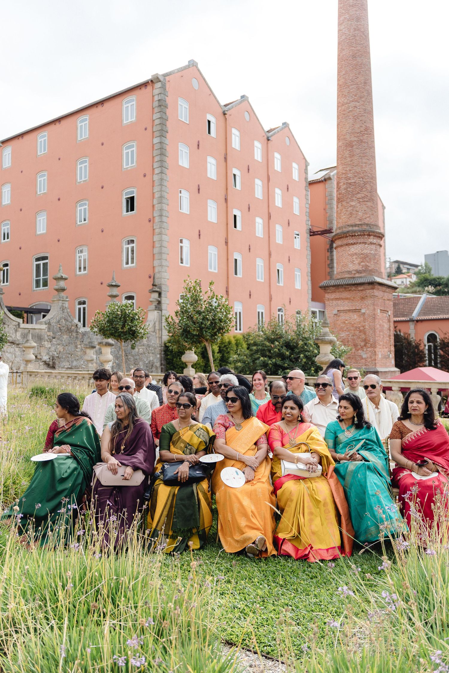 Wedding guests in colorful traditional attire sit together on grass near brick buildings.