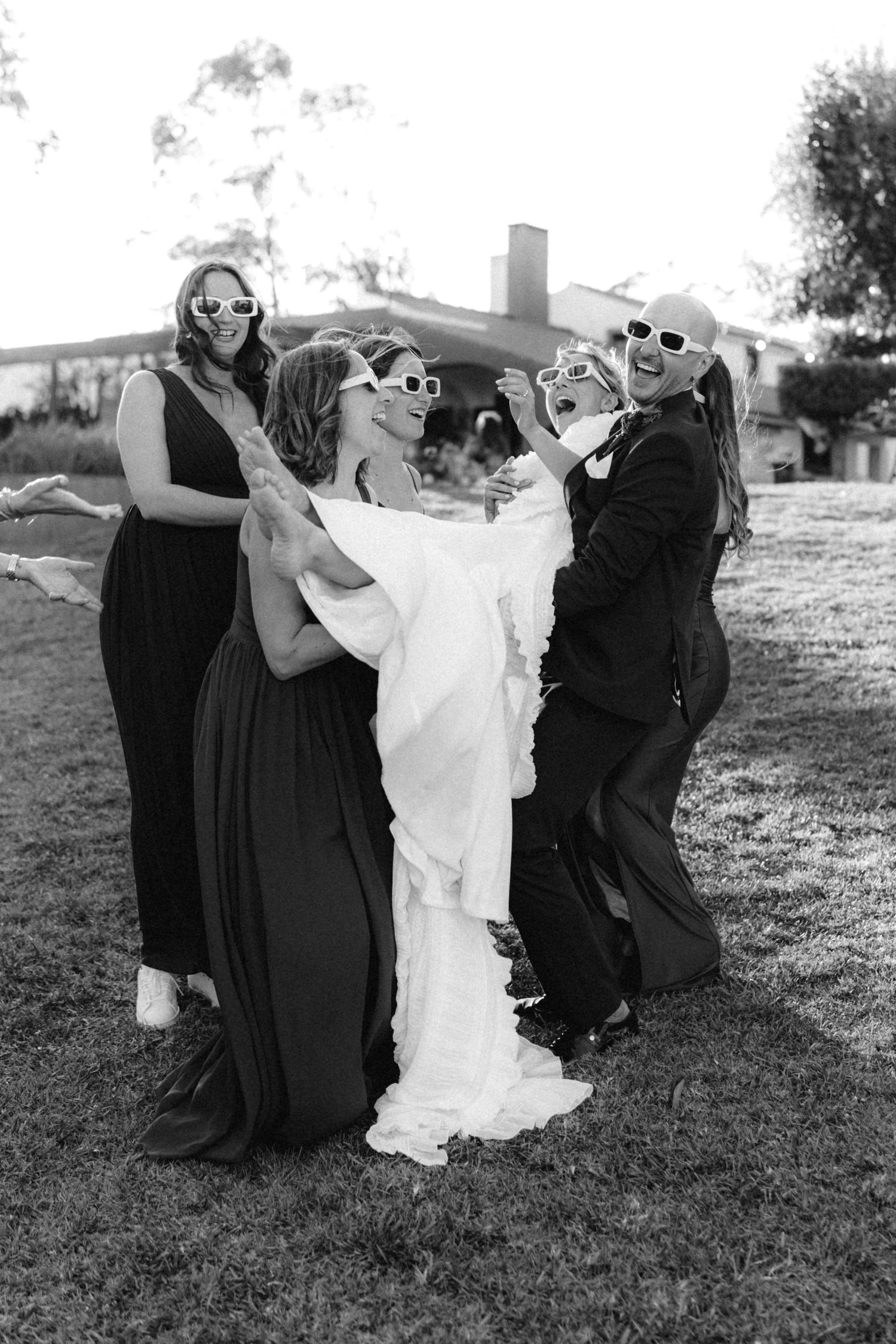 Wedding party in black dresses playfully dips and poses together on a grassy hill.