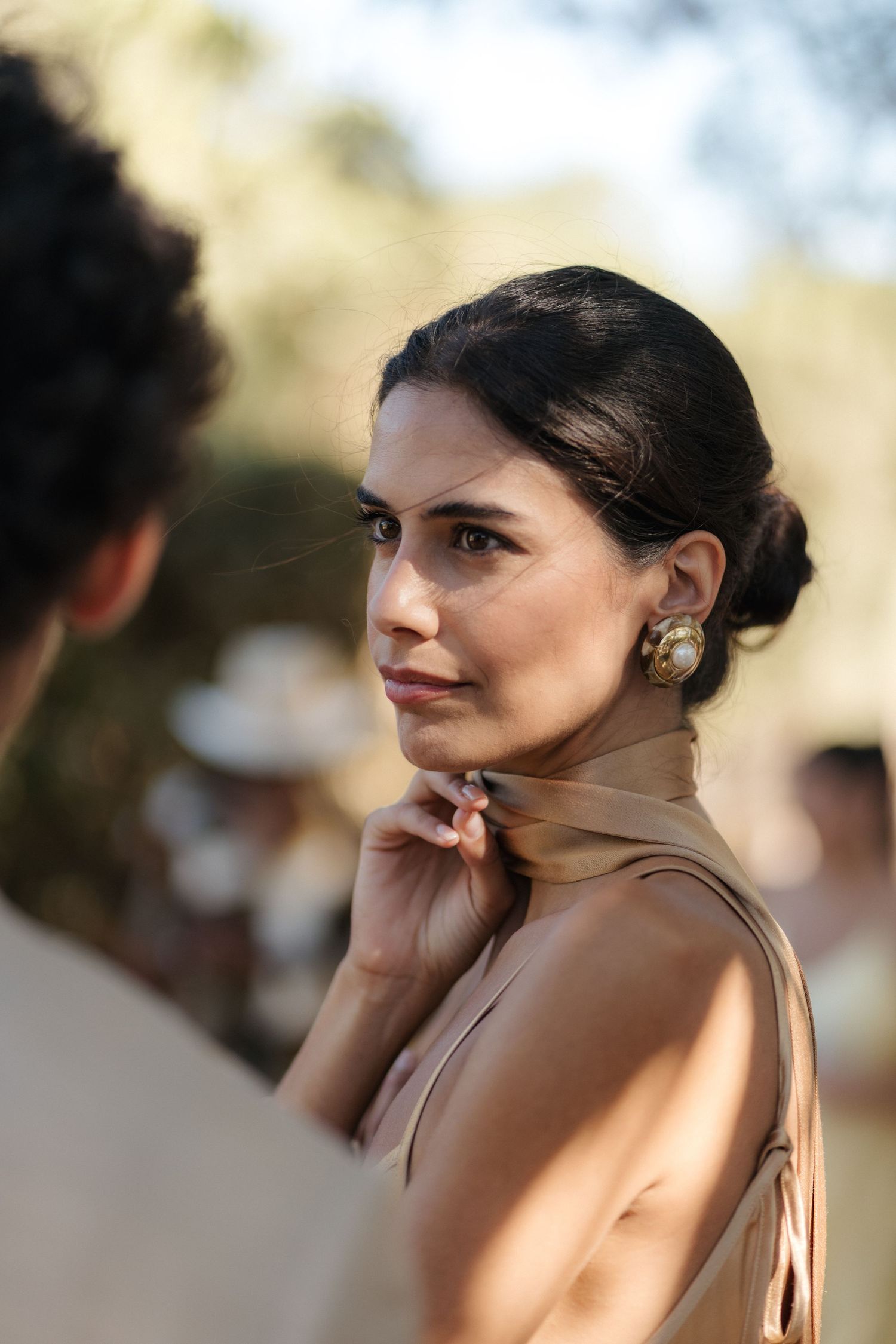 Guest in elegant beige dress with classic updo at outdoor celebration.