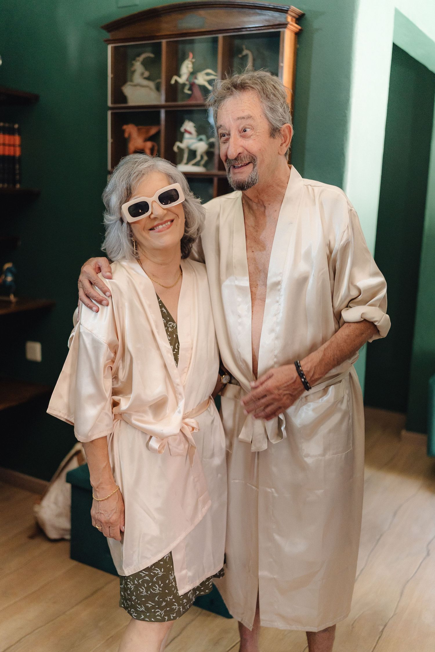 Couple in matching robes posing together against green wall with display cabinet.