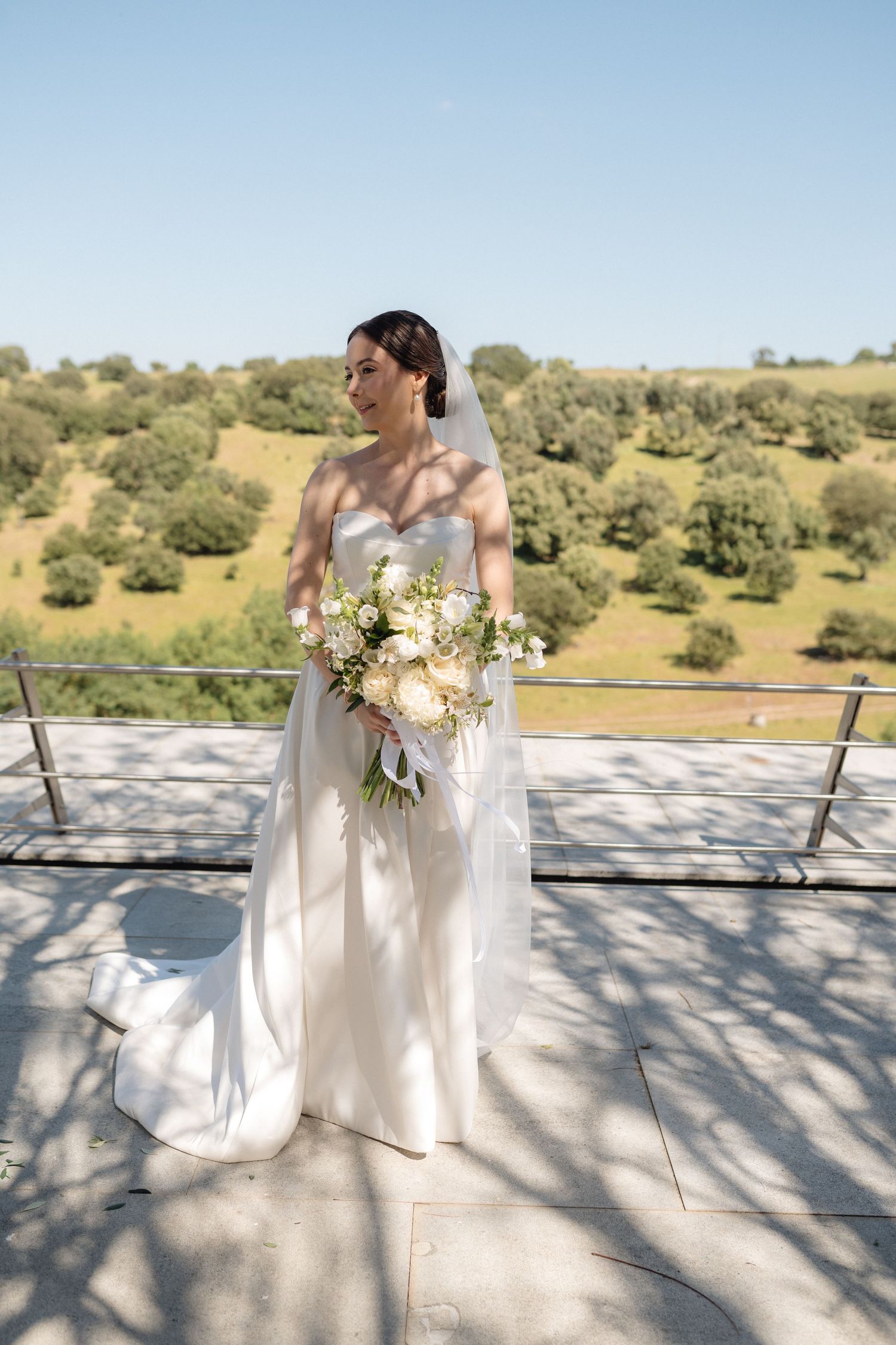 A bride in an elegant strapless gown holds a white bouquet on an outdoor terrace with rolling hills behind.
