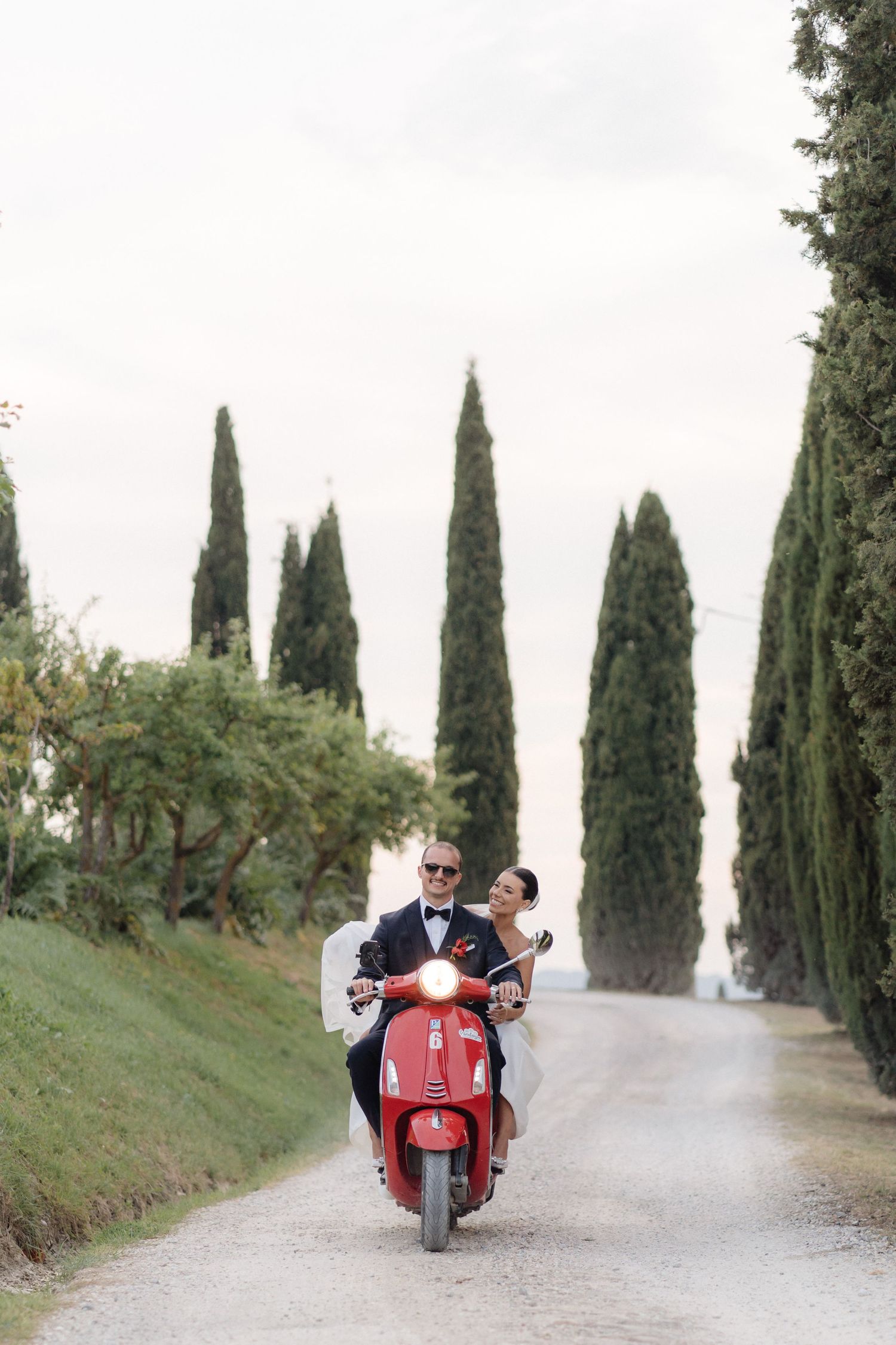 Bride and groom riding a red Vespa down a scenic cypress-lined avenue in Tuscany.