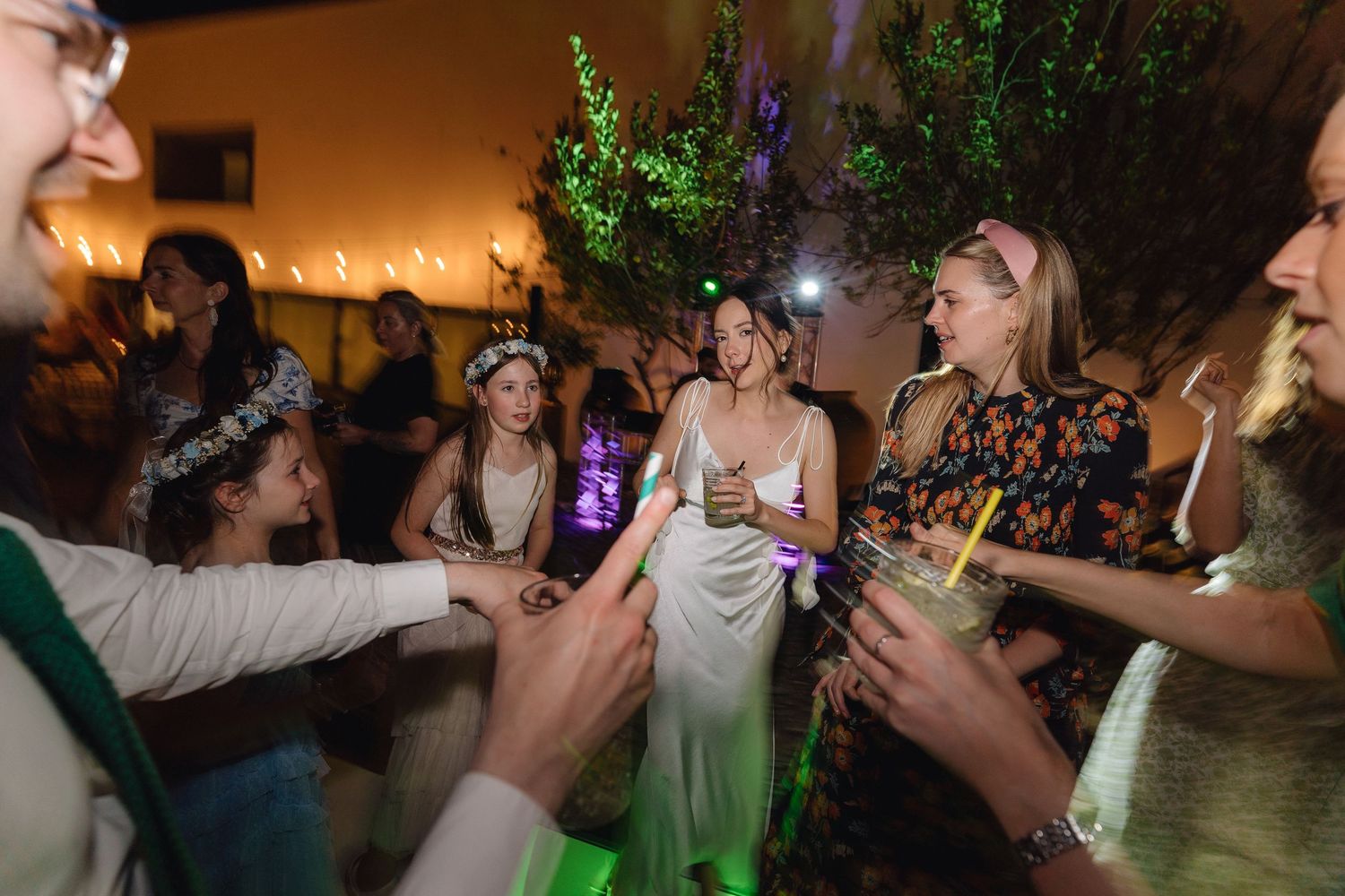 Festive crowd dancing beneath decorative greenery and warm lighting at an indoor celebration.