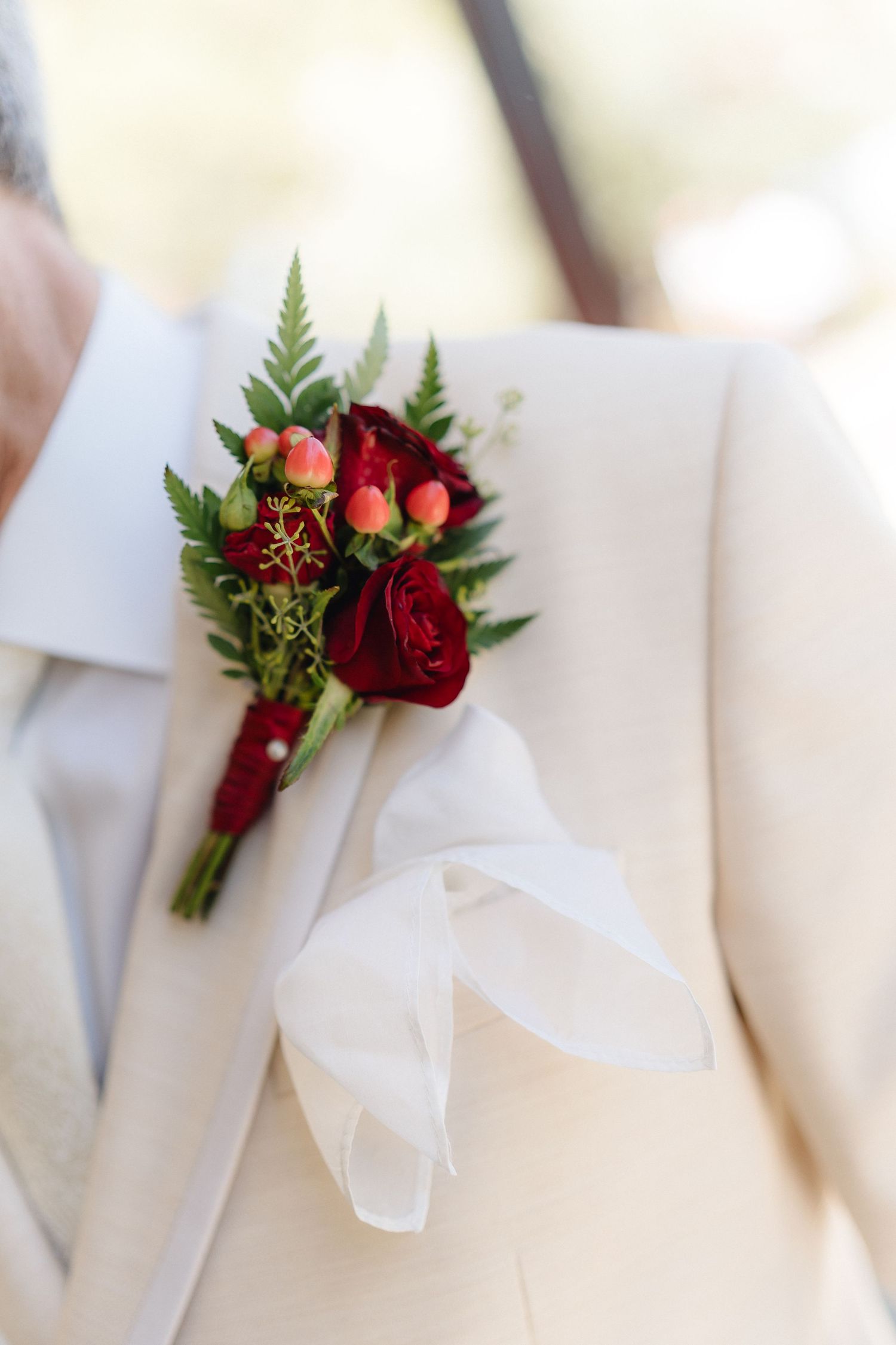 Deep red and pink floral boutonniere with ferns pinned to white suit lapel.