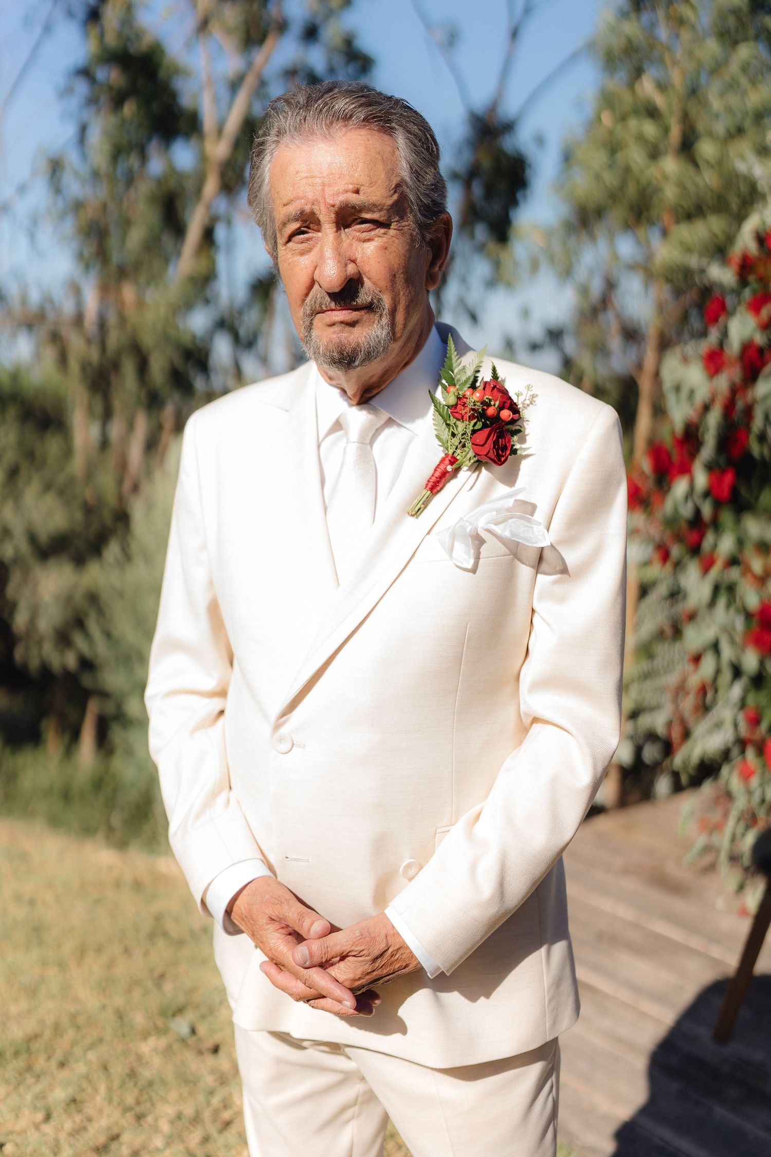 A person in a cream-colored suit with red boutonniere poses outdoors.