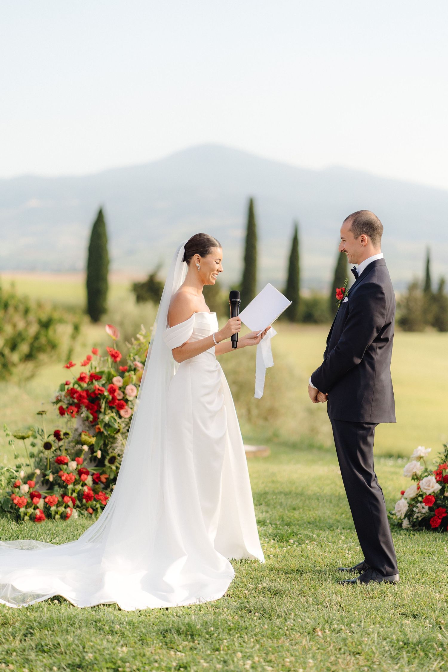 Bride reading vows to groom during outdoor ceremony with cypress trees and mountains backdrop.