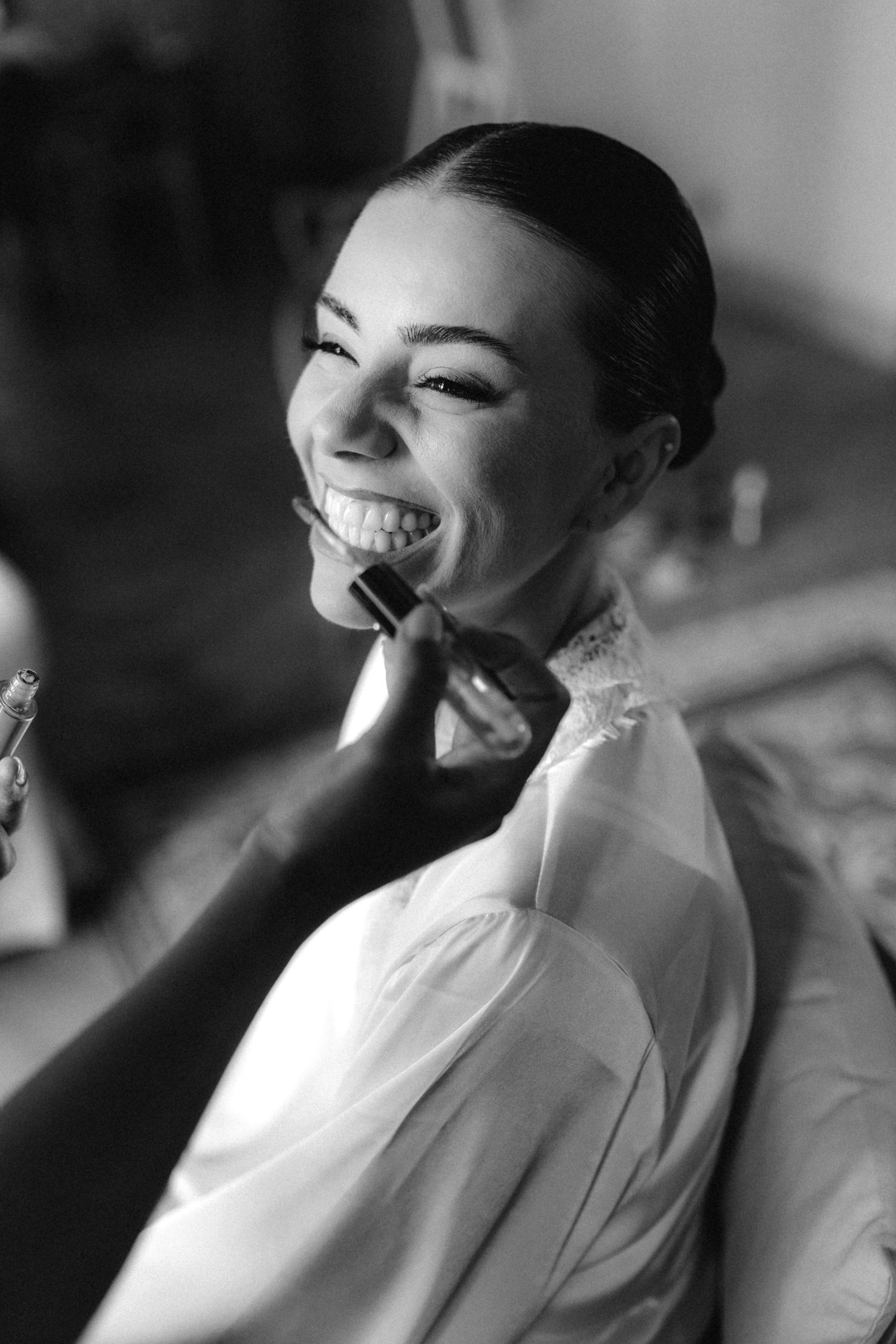 Black and white photo of a smiling bride getting makeup applied with a brush on her wedding day.