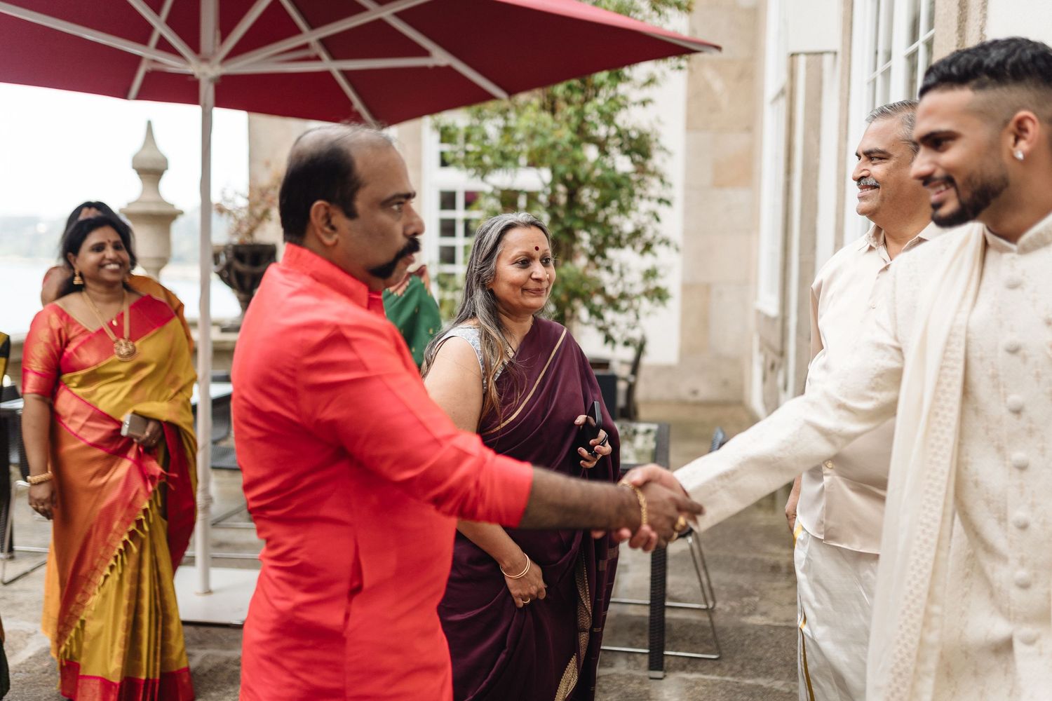 Wedding guests in traditional attire greeting each other under red umbrella outdoors.