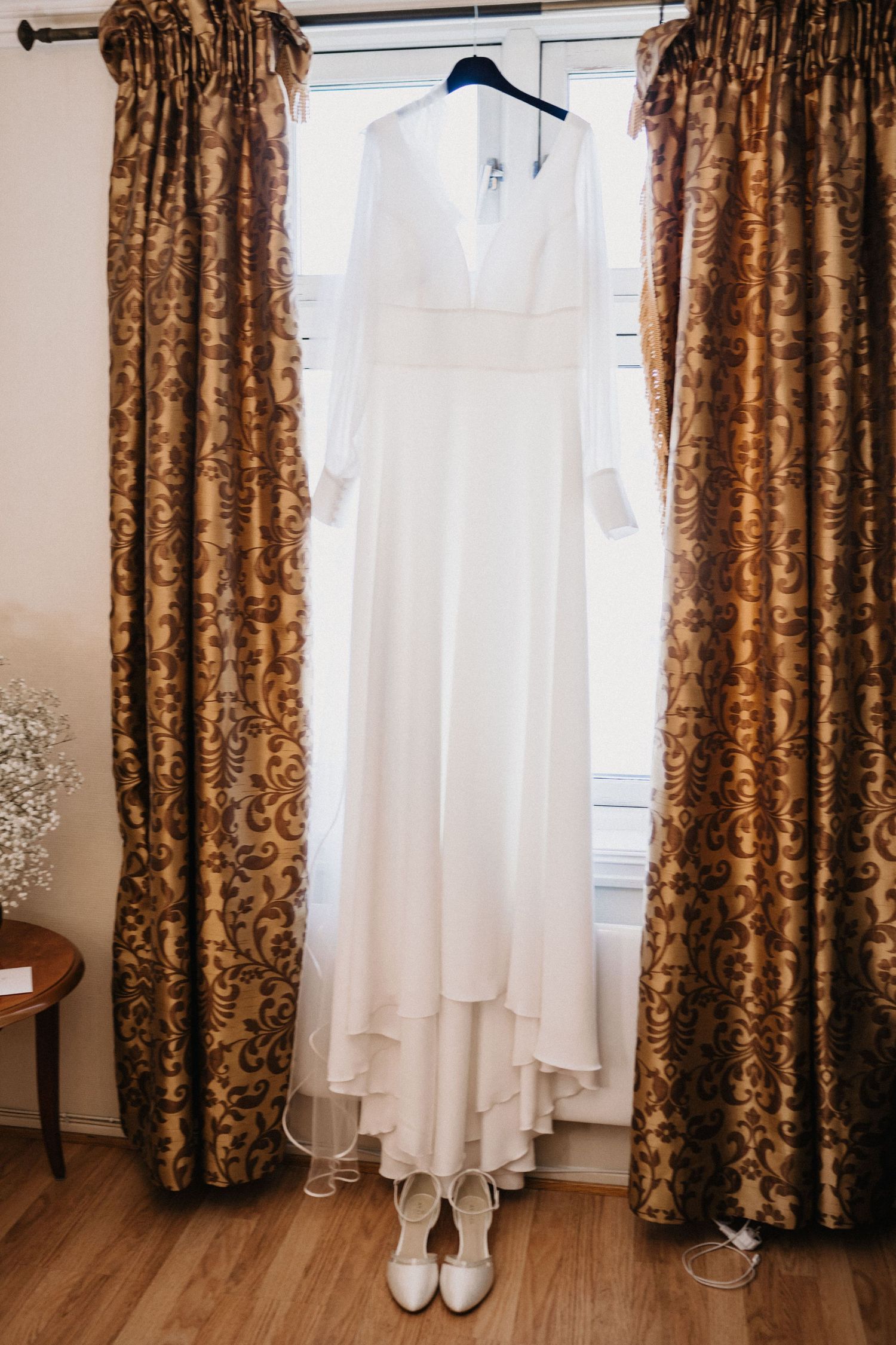 A row of wedding details including a white dress, brown floral curtains, and bridal shoes displayed in a room.