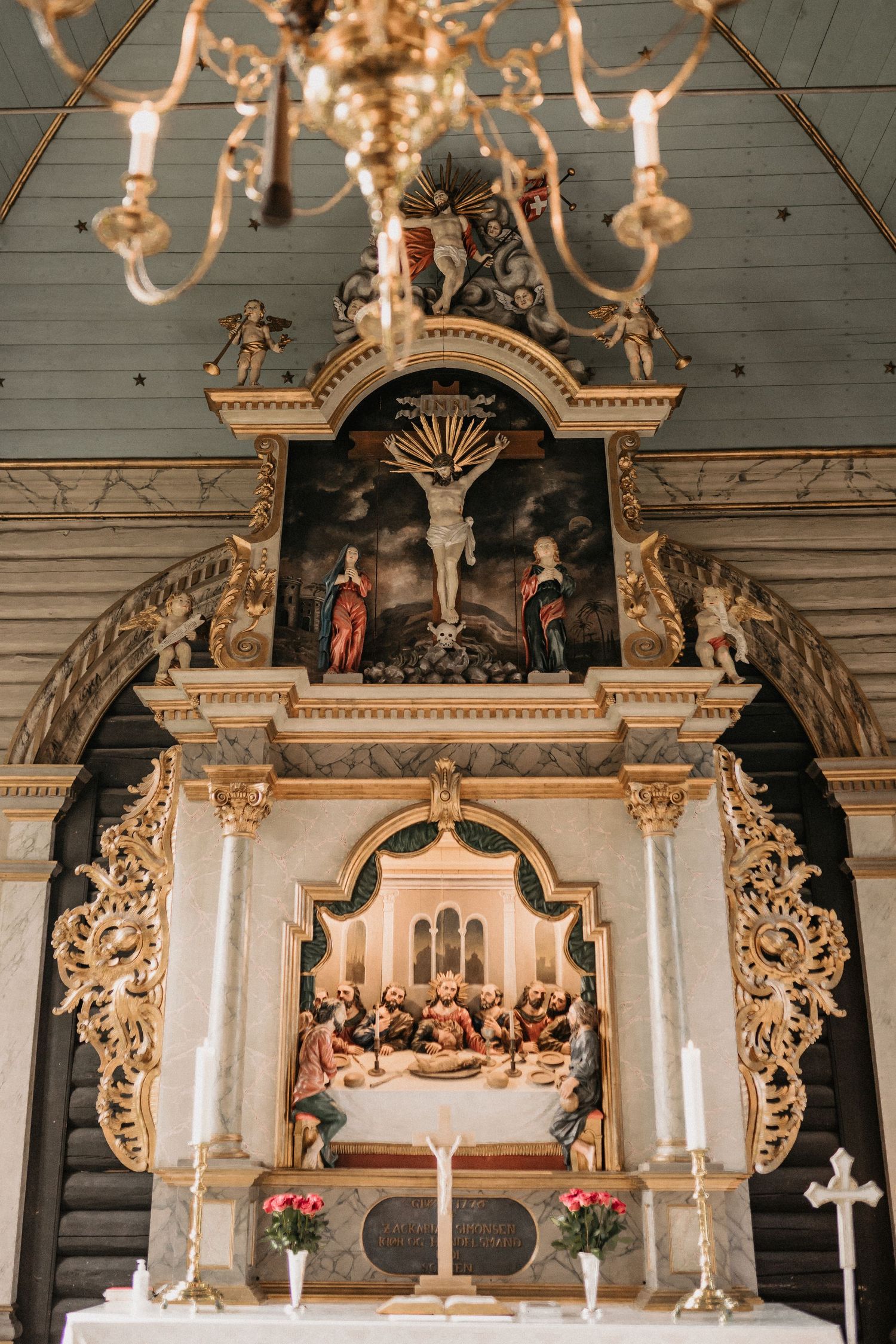 Ornate church altar with religious artwork, golden details, and a crucifix centerpiece illuminated by chandelier light.