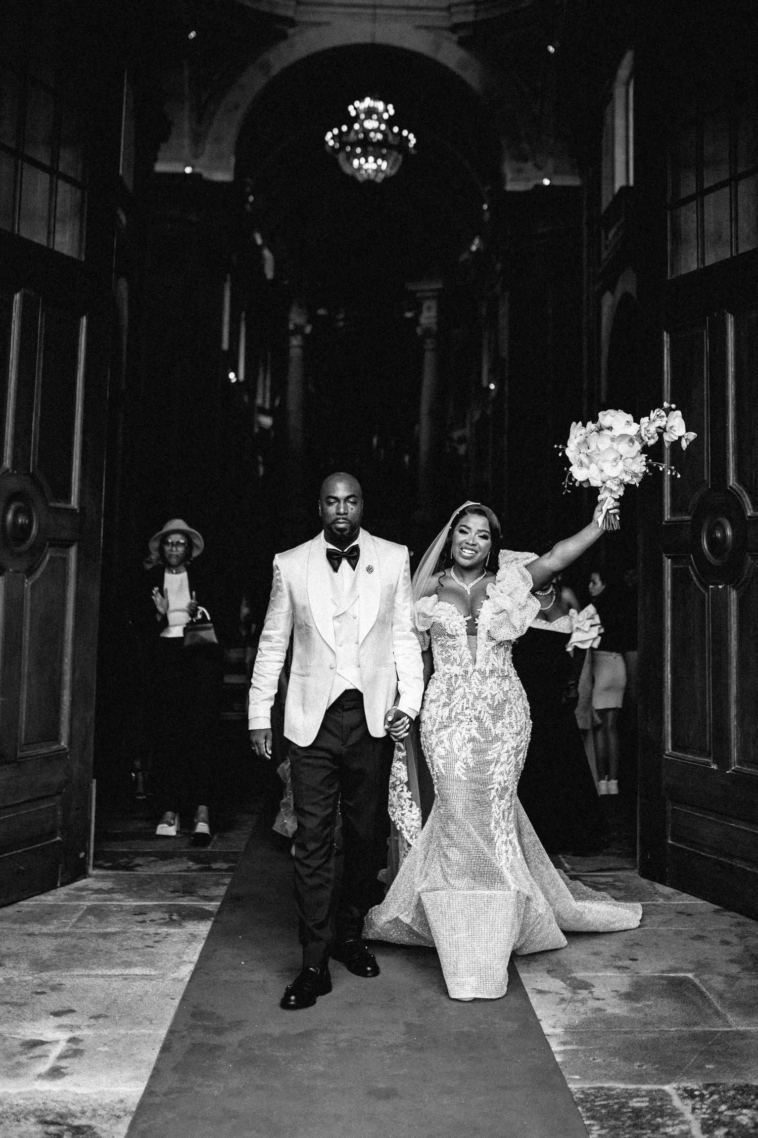 A glamorous wedding couple exits a grand church venue with the bride holding a bouquet in a stunning mermaid gown.