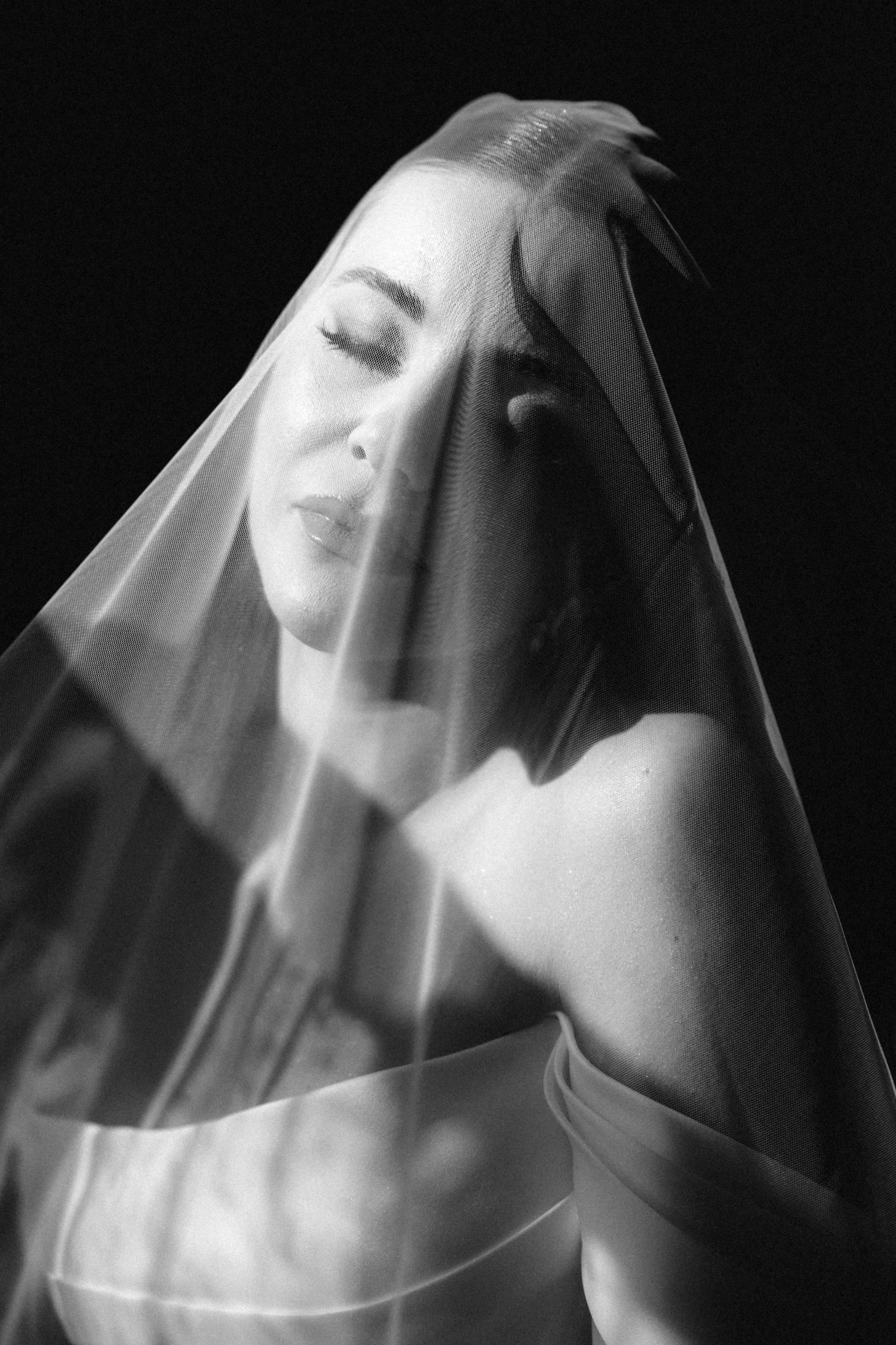 Black and white bridal portrait of a woman with closed eyes draped in a sheer wedding veil against dark background.