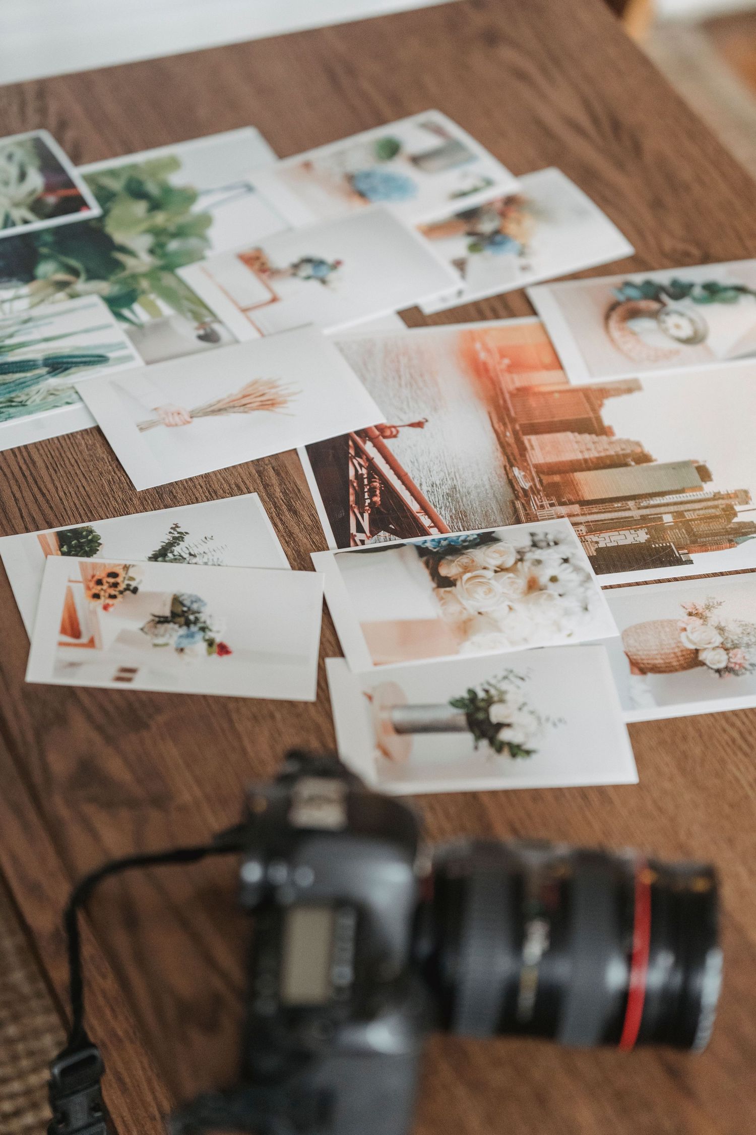A DSLR camera sits on a wooden table surrounded by scattered printed photos showcasing various lifestyle and nature subjects.