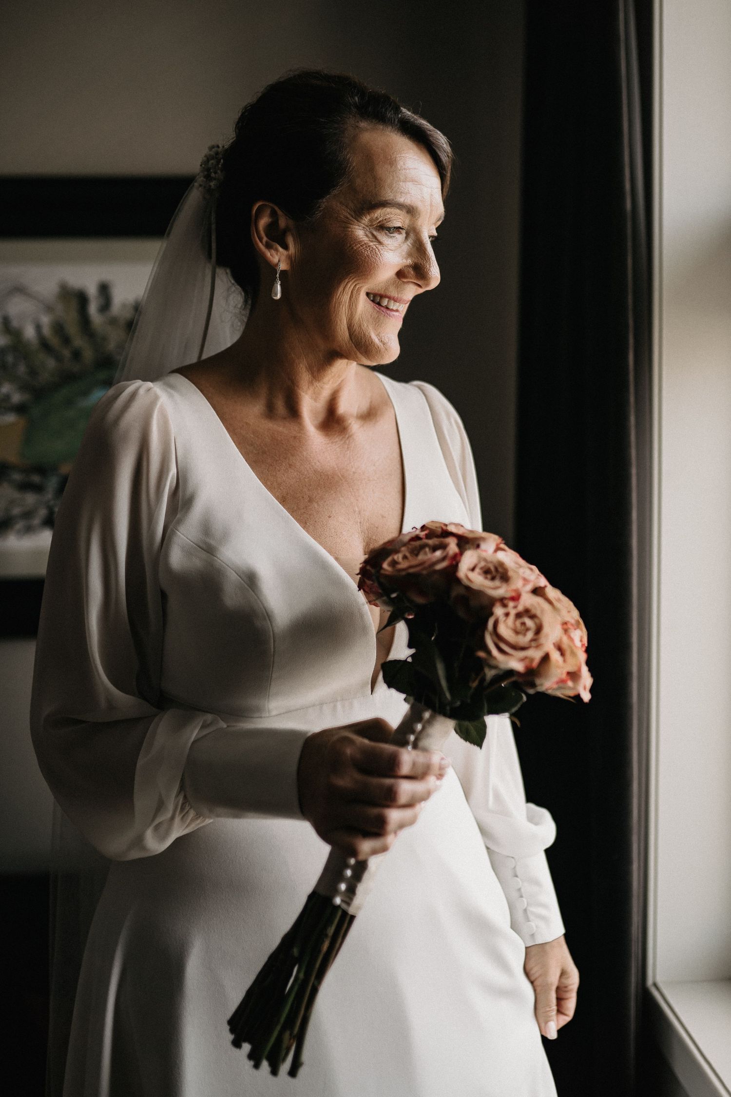 Elegant bride in long-sleeved white dress holding bouquet near window in moody lighting.
