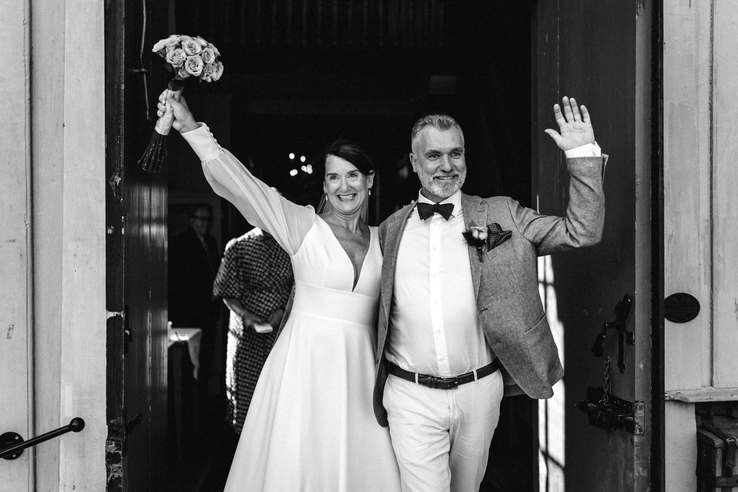 Wedding party exiting venue with celebratory gestures in black and white photography.