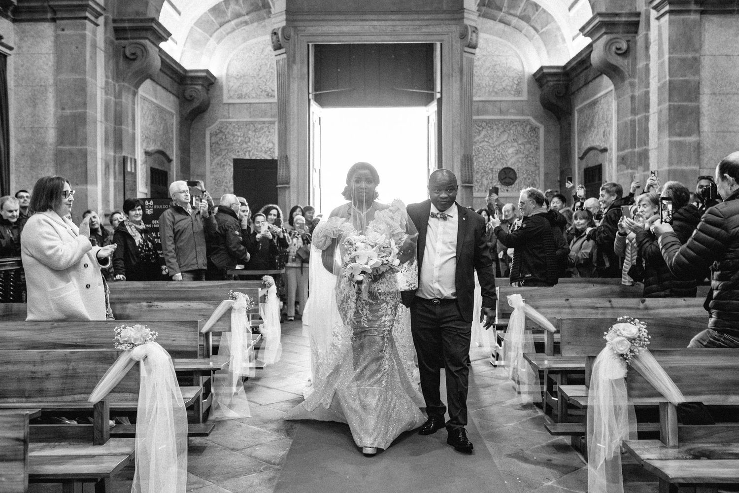 Black and white photo of wedding recessional in grand church with guests looking on.