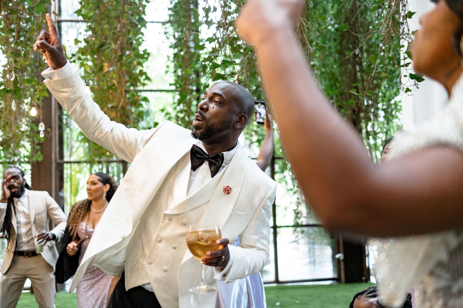 Wedding celebration with guests dancing and raising hands under green vine decorations.