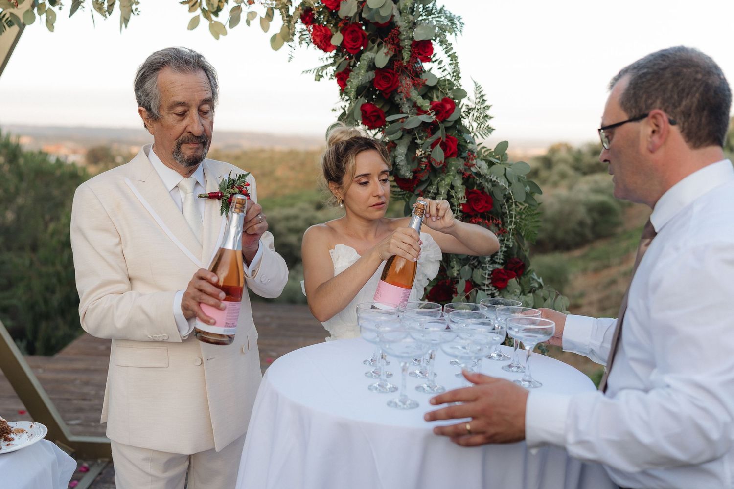 Wedding celebration toast under arch decorated with red roses and greenery.