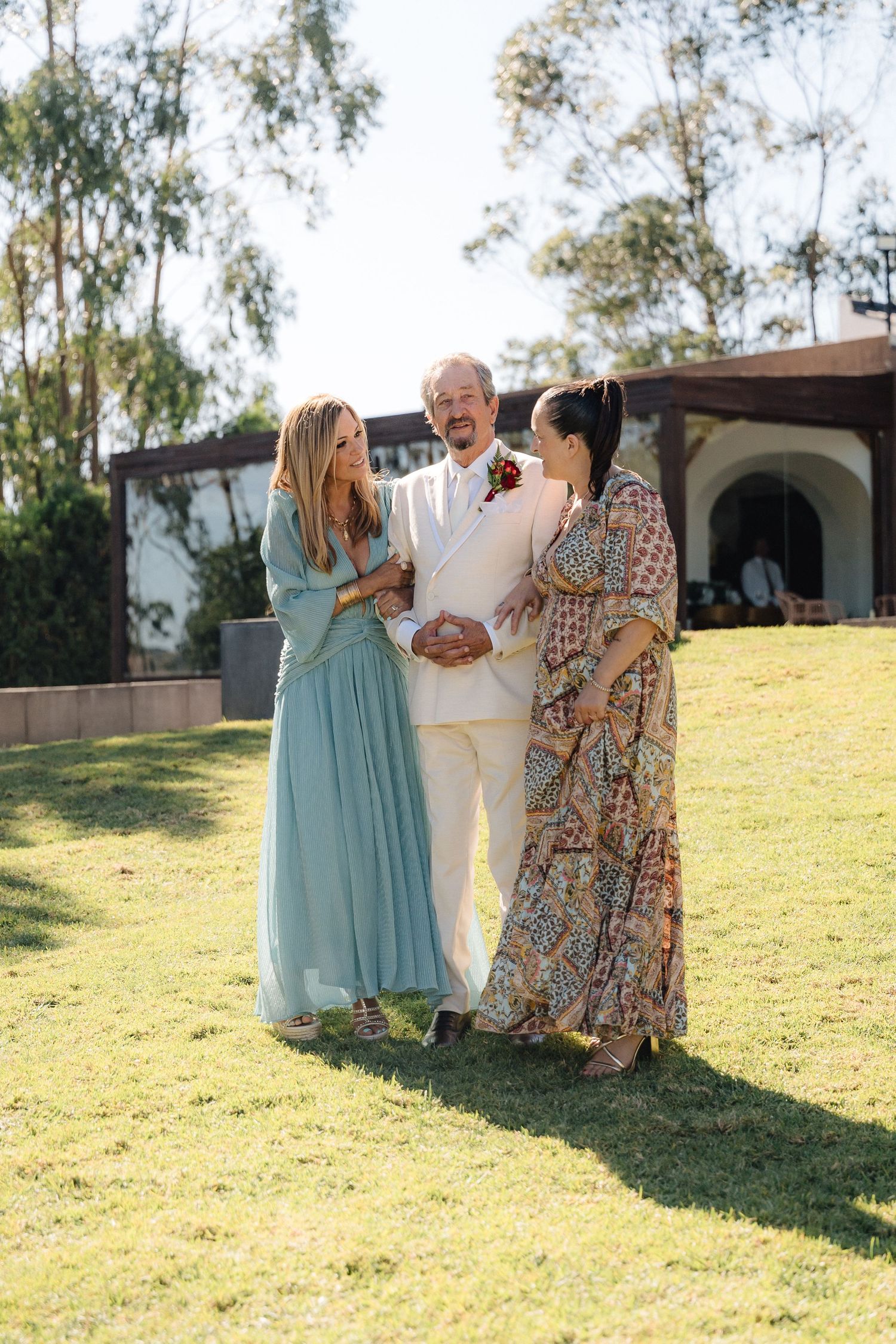 Wedding guests in elegant attire interact on a sunny grassy hillside.