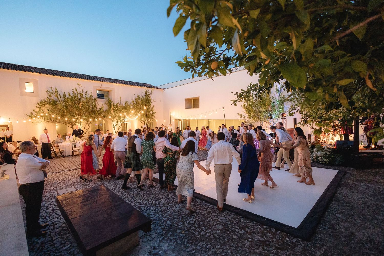 Panoramic view of an outdoor wedding reception with string lights and white buildings.