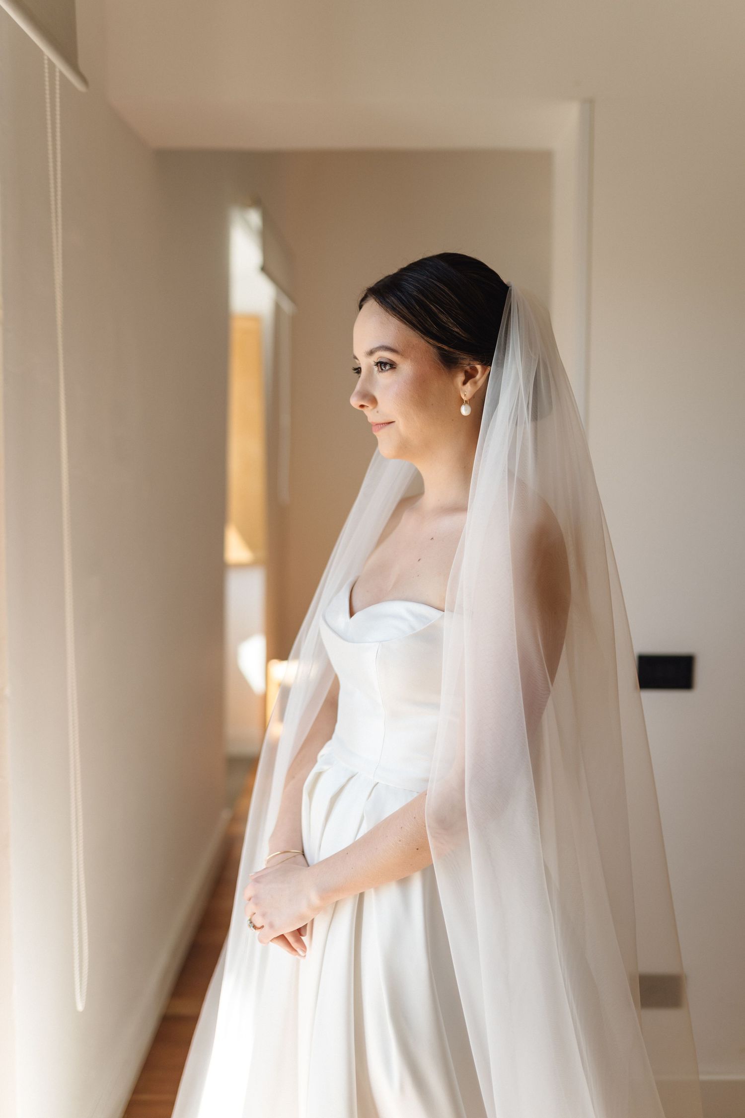 Side profile of bride in white wedding gown standing in naturally lit corridor.